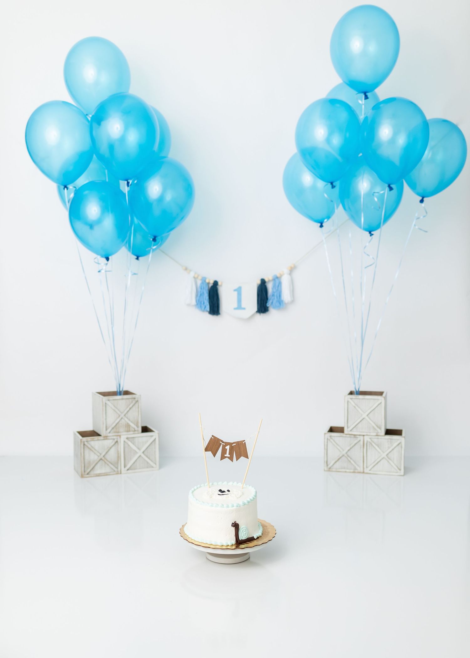 Blue balloons and white boxes arranged around white cake for cake smash photography setup.