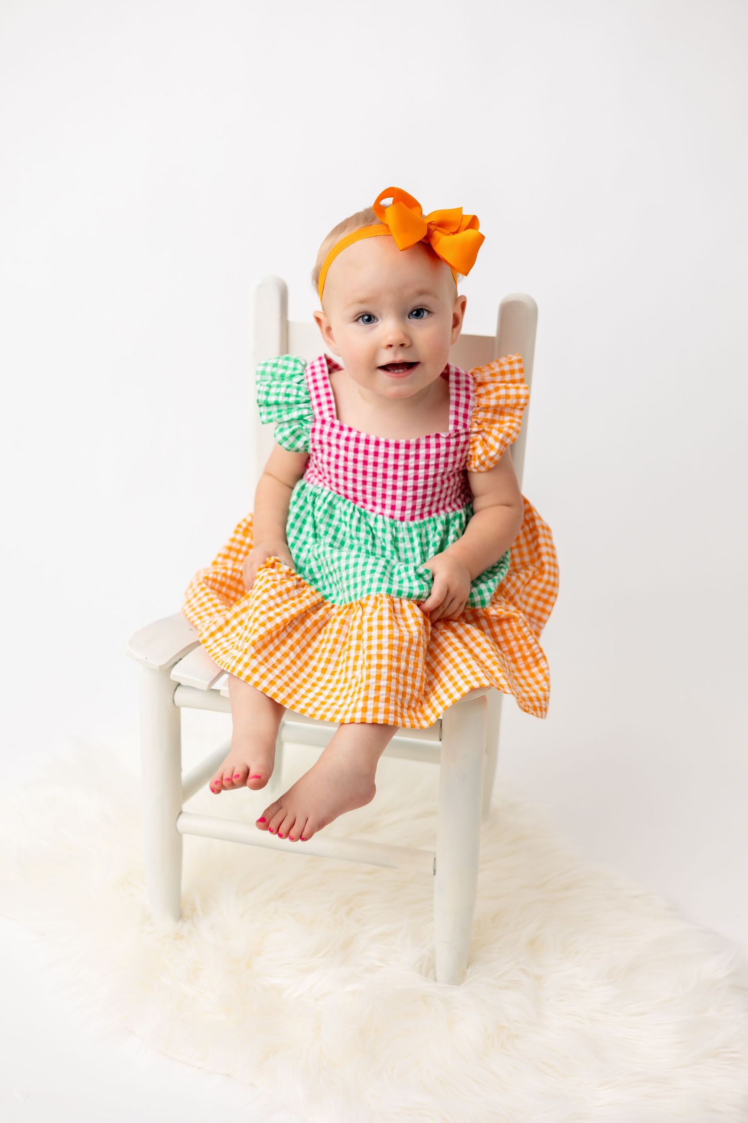 Baby girl in colorful dress with ruffles sitting on white chair against light background for her one year portrait.