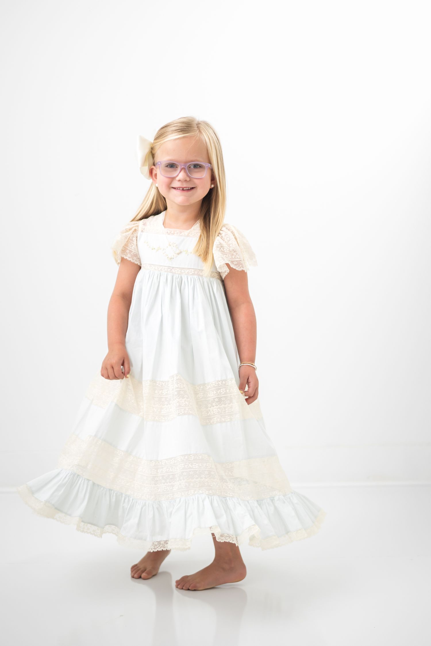 Young blonde girl in white lace dress and glasses smiling barefoot in studio.