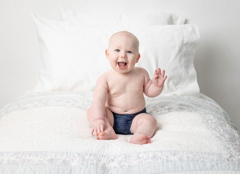 Baby boy celebrating 6 months old wearing navy shorts waves while sitting on white bedding.
