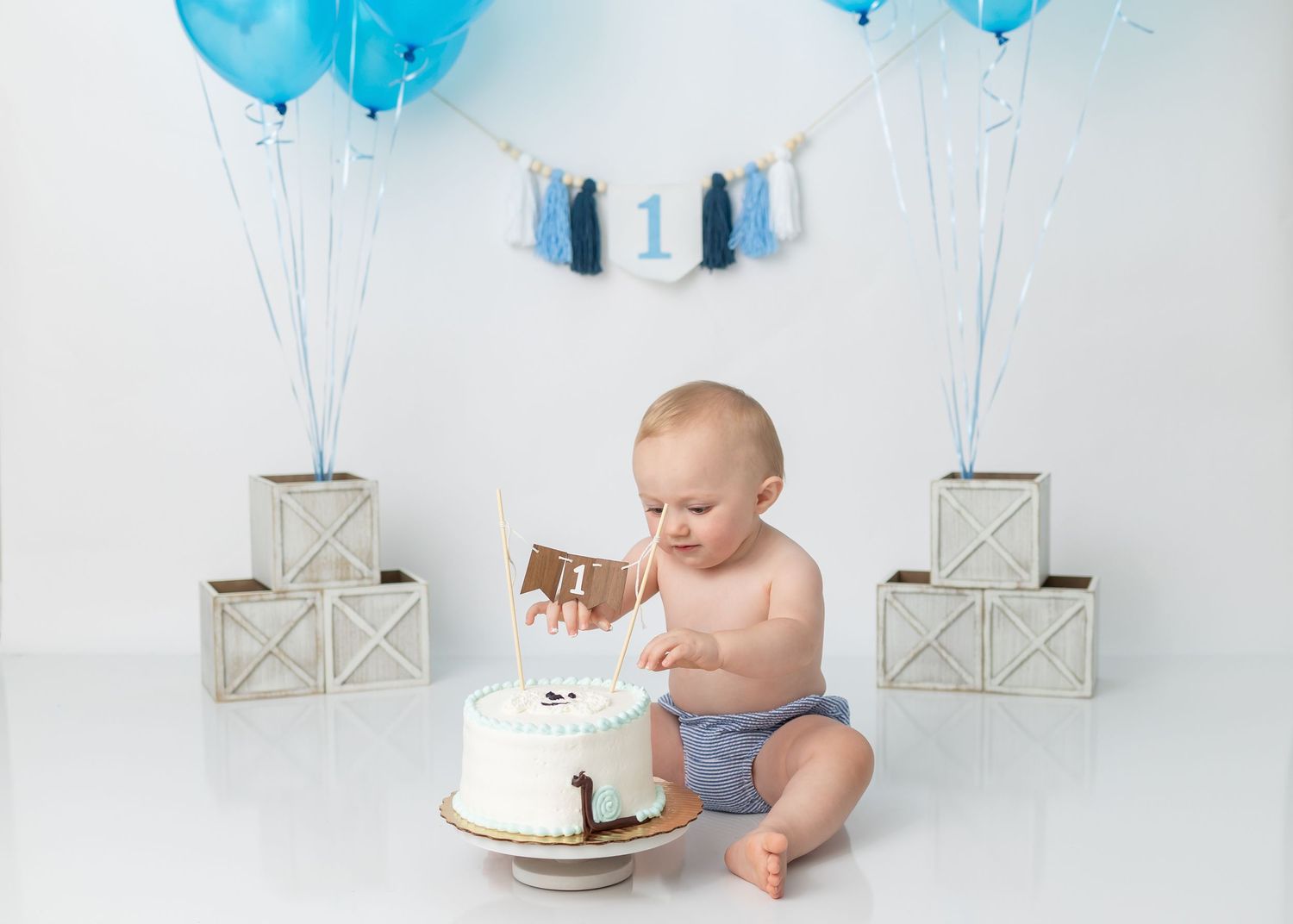 Baby in diaper reaching for white birthday cake surrounded by blue balloons and decorative boxes.