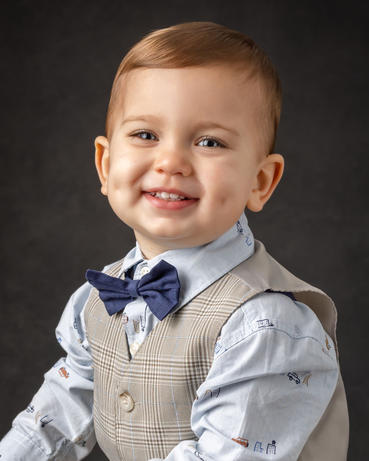 A baby in formal attire with a blue bow tie and tan plaid vest smiles brightly against a dark background.