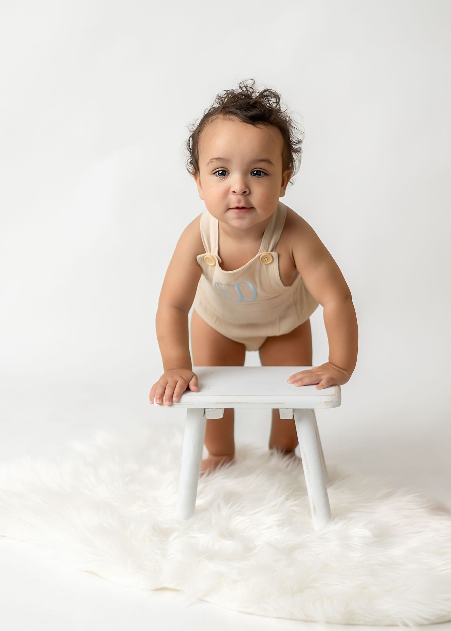 Baby in cream outfit poses on small white chair against white background for one year portrait session.