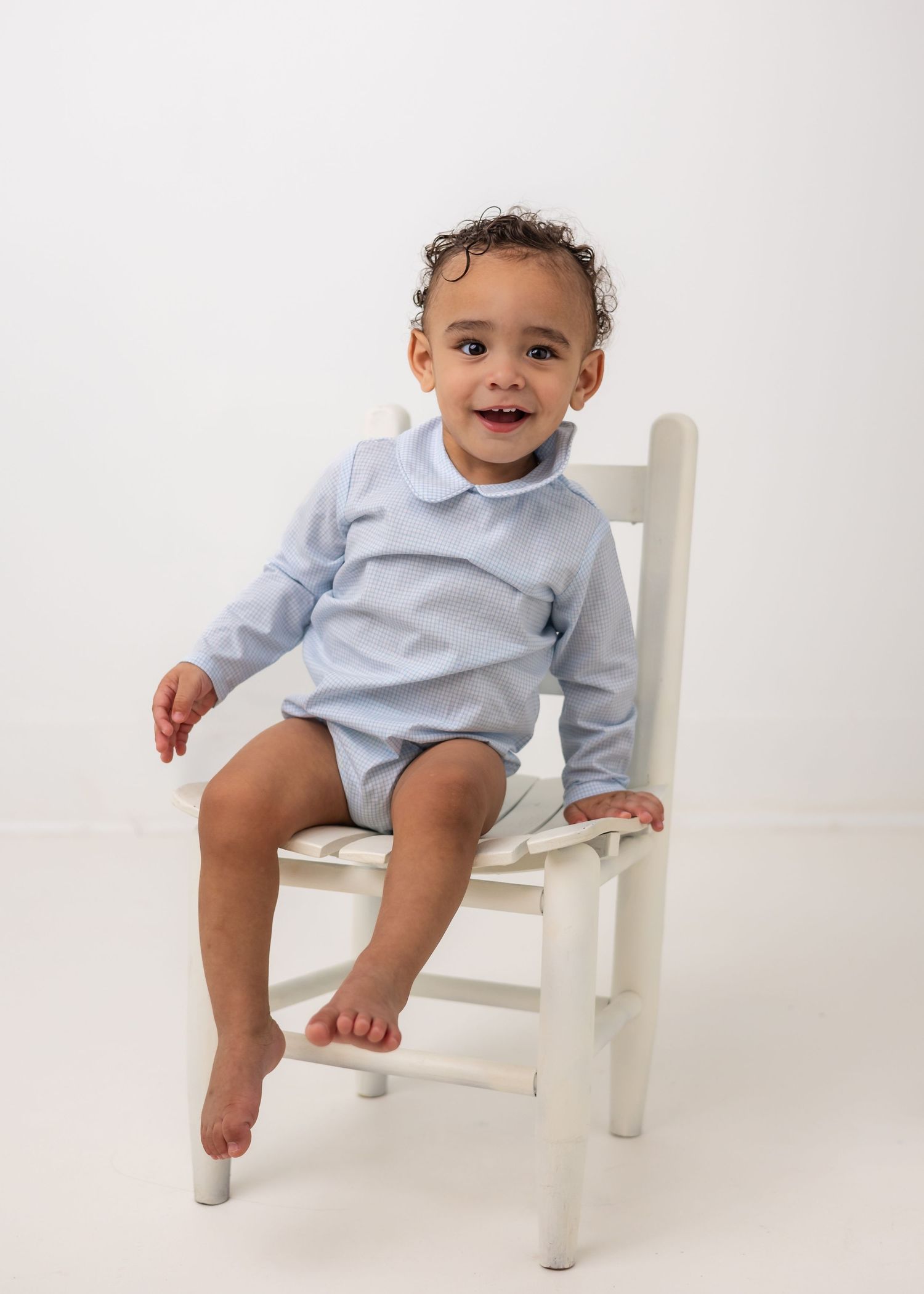 Child in light blue outfit sitting on white wooden chair against white background.