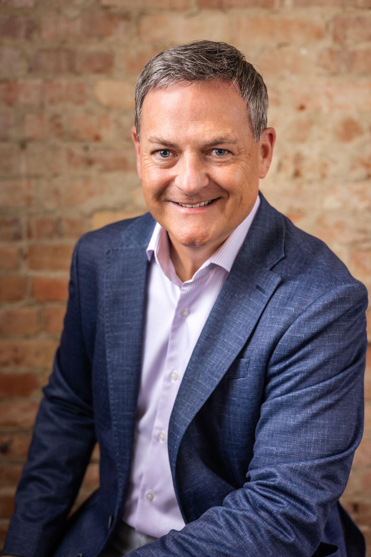 Professional headshot series of someone in a navy suit and pink shirt against a rustic brick wall background.