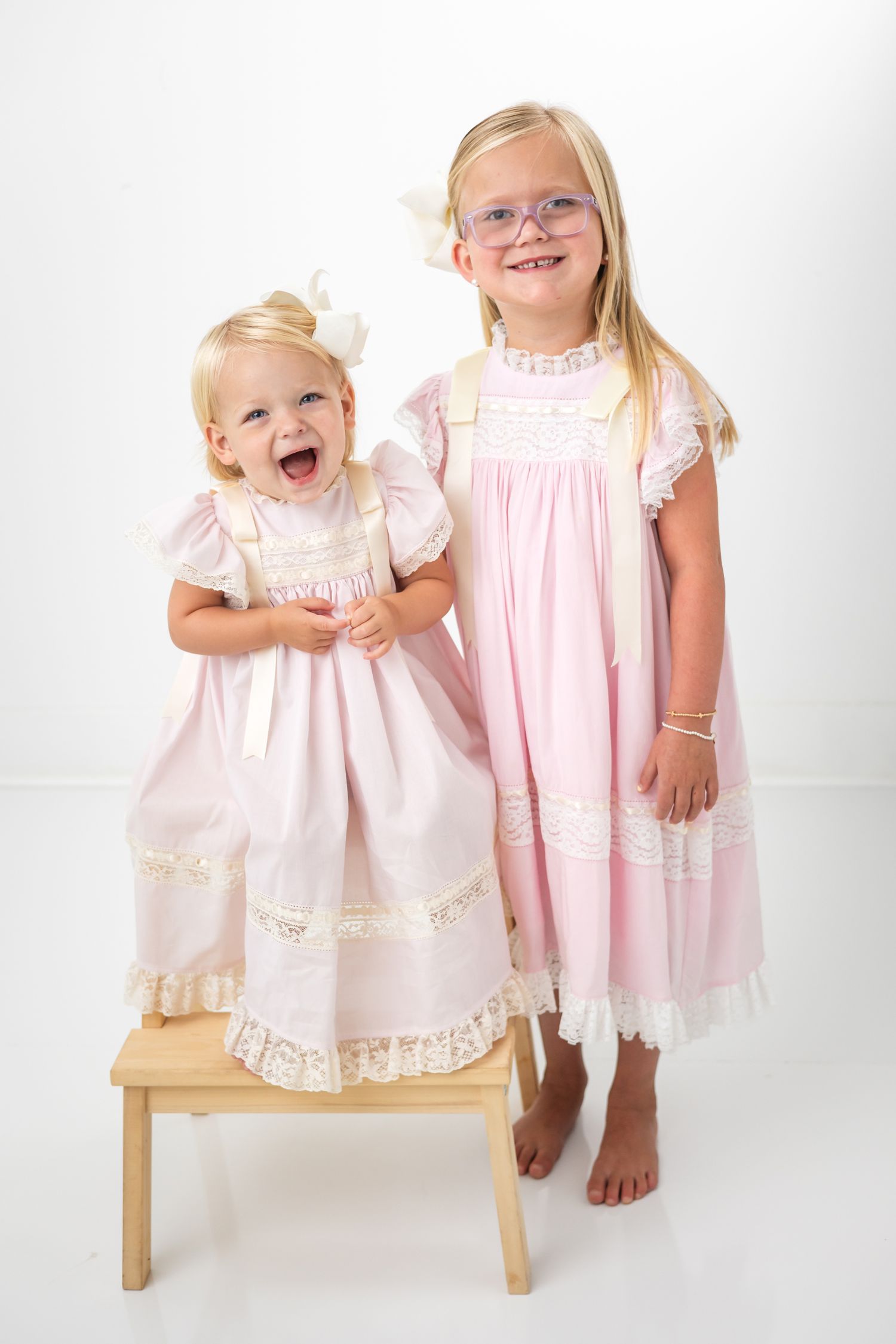 Two young girls in matching pink lace dresses, one wearing glasses, posing together on a wooden step stool.
