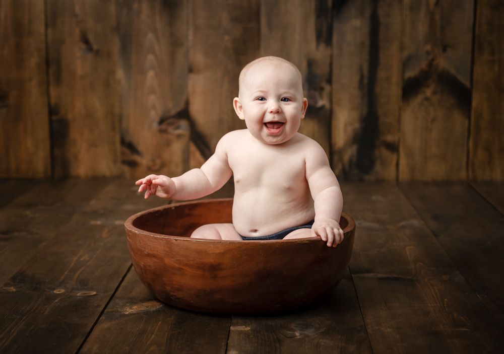 Smiling baby sits in large wooden bowl against rustic wooden wall background.