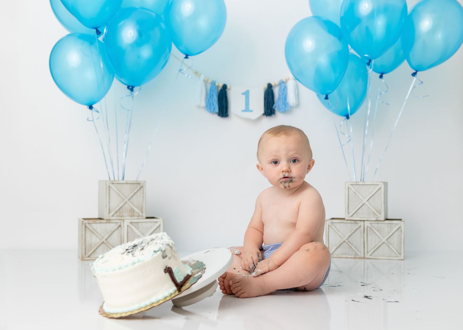 Baby sitting beside messy smashed cake with blue balloon decorations.
