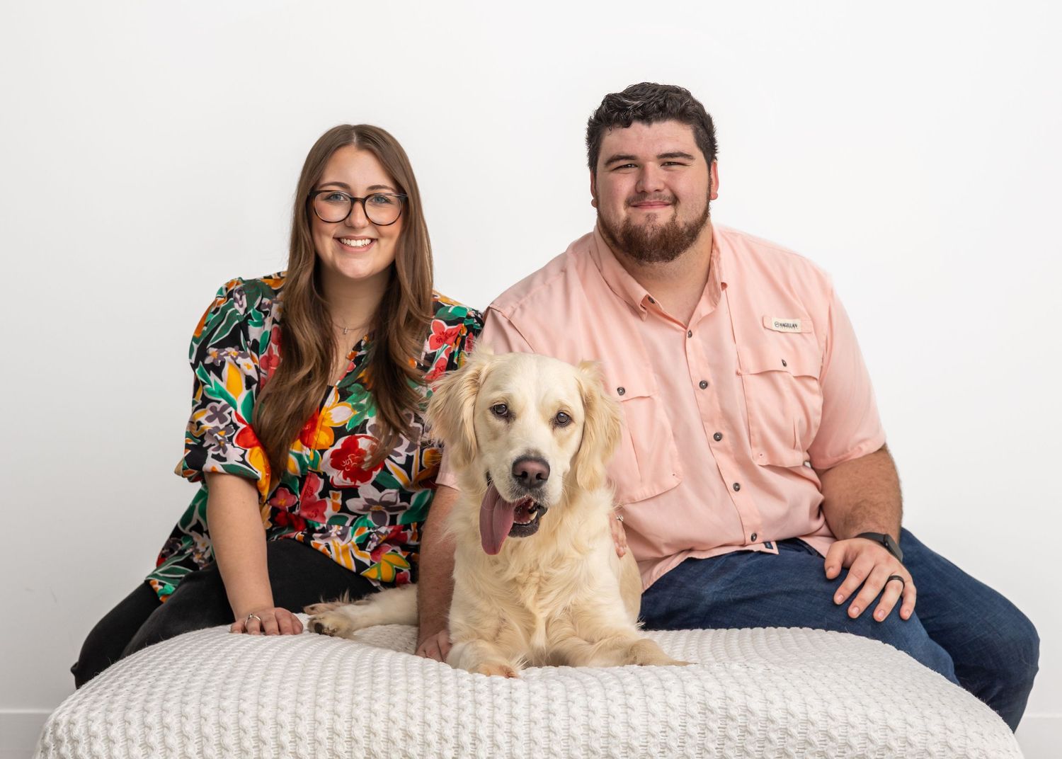 A Golden Retriever sits between two people on a white cushioned bench in a studio portrait setting.
