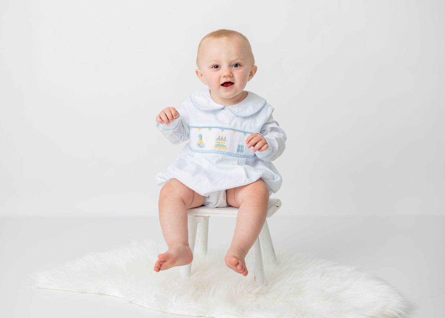 Baby in blue outfit sitting on small white stool against light background.