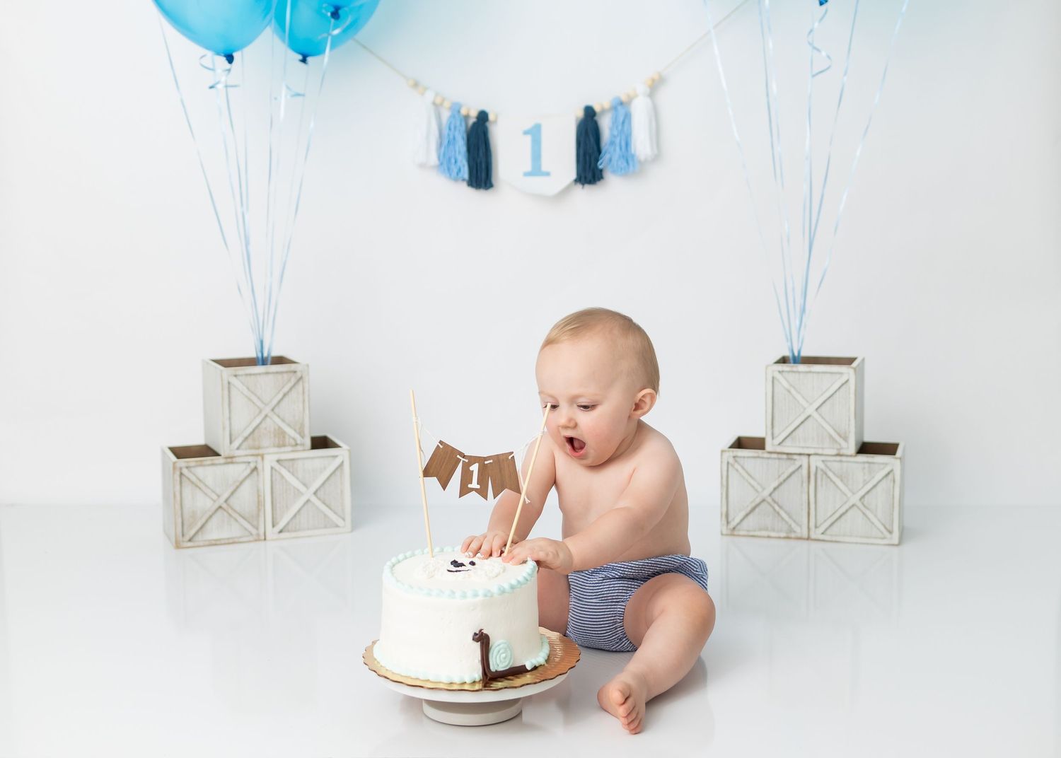 Baby exploring birthday cake surrounded by blue balloons and white boxes.