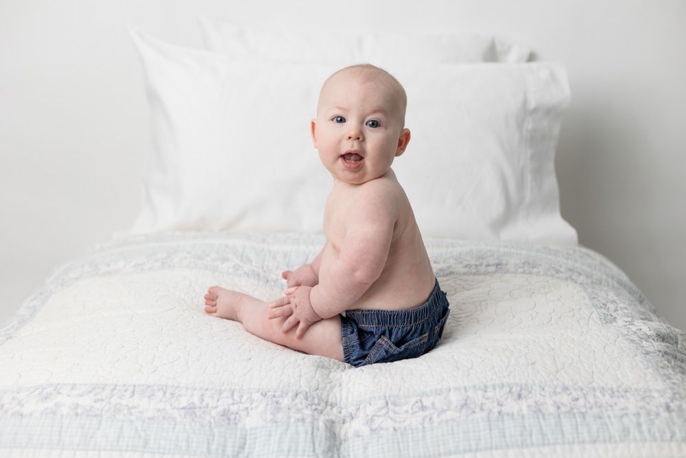 Baby in dark shorts sits on white bed with pillows in background.