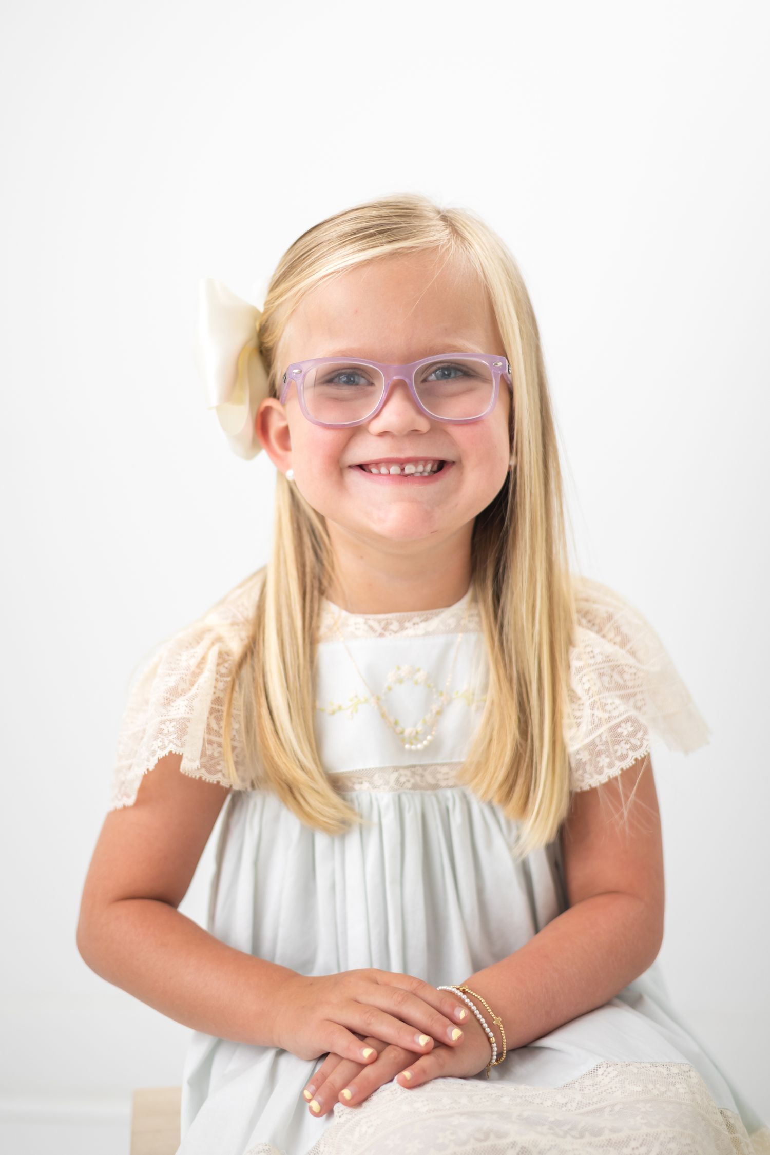 Smiling young blonde girl wearing pink glasses and a white lace dress with a bow in her hair.