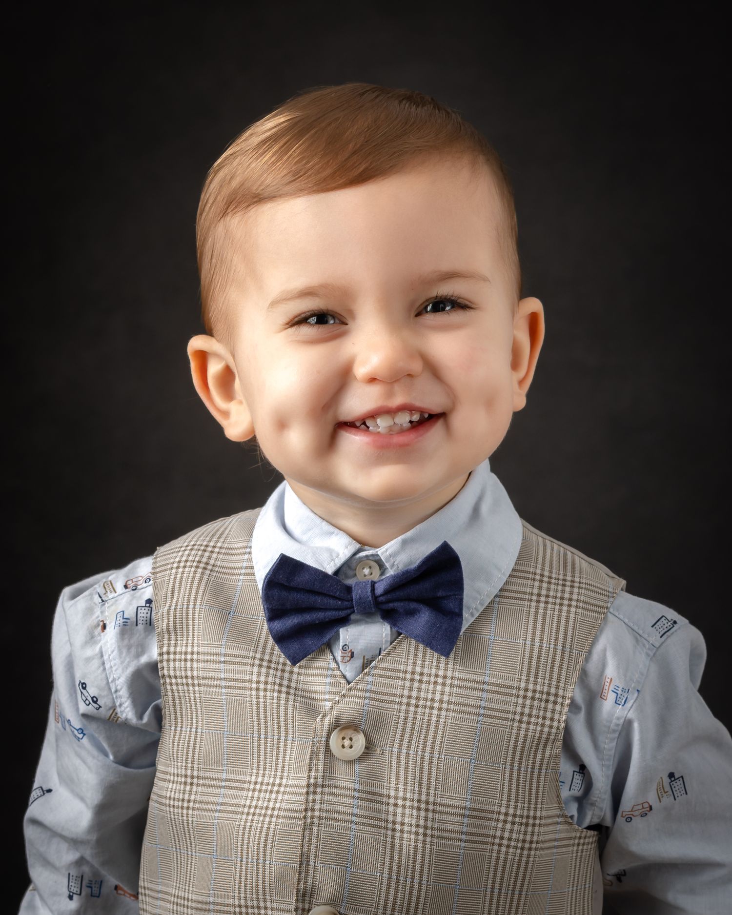 A small child wearing a plaid vest and navy bowtie smiles warmly against a dark background.