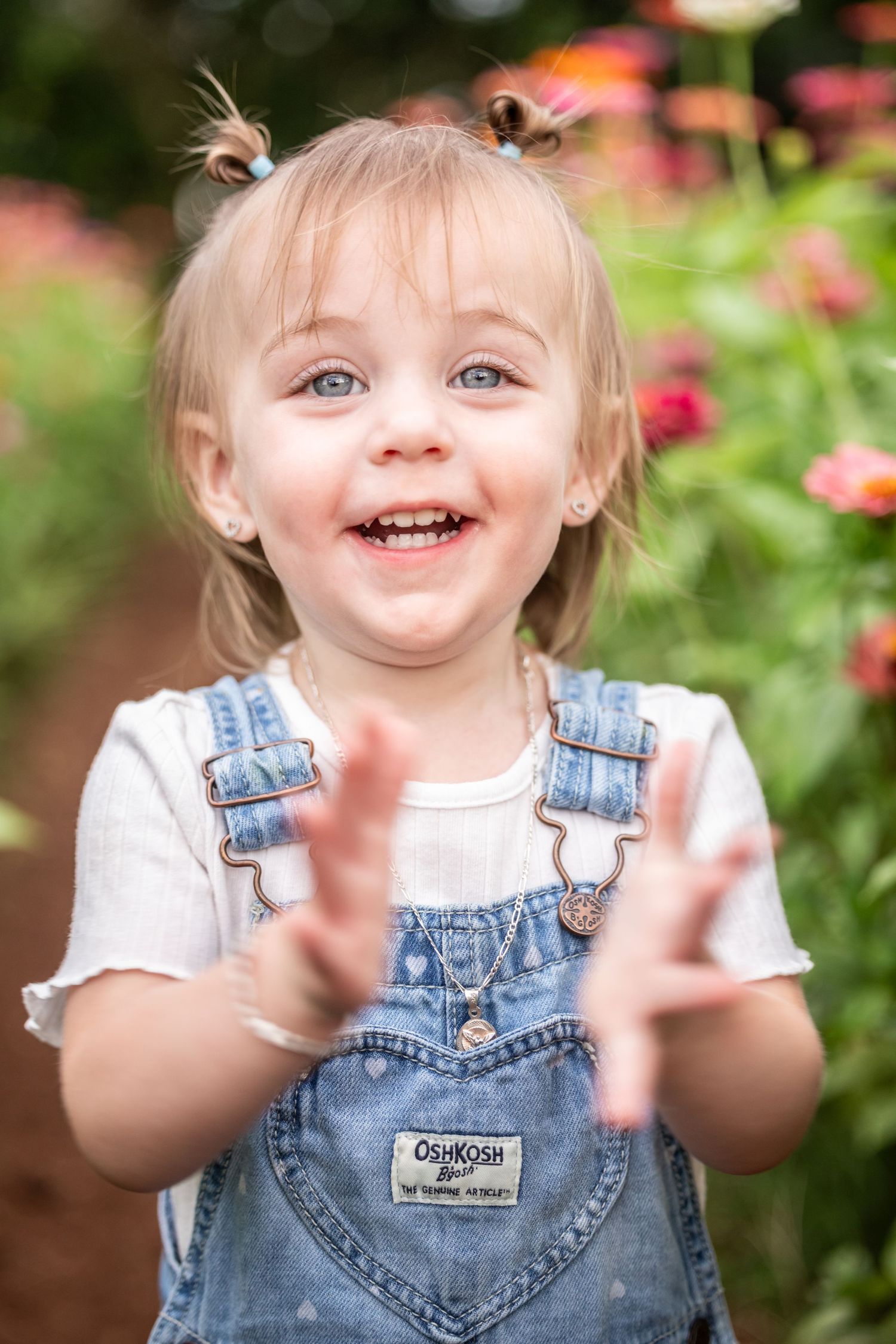 Smiling toddler girl in denim OshKosh overalls clapping hands in a colorful flower garden just outside of Enterprise, AL