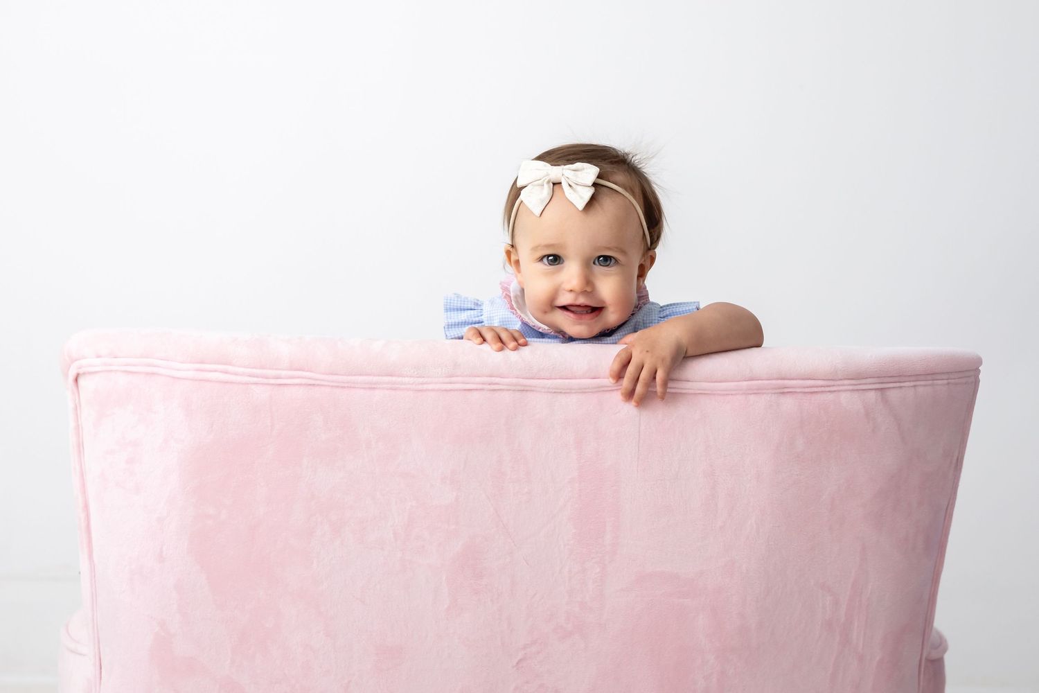 Smiling baby girl with bow headband peeking over a pink velvet chair against a white background.