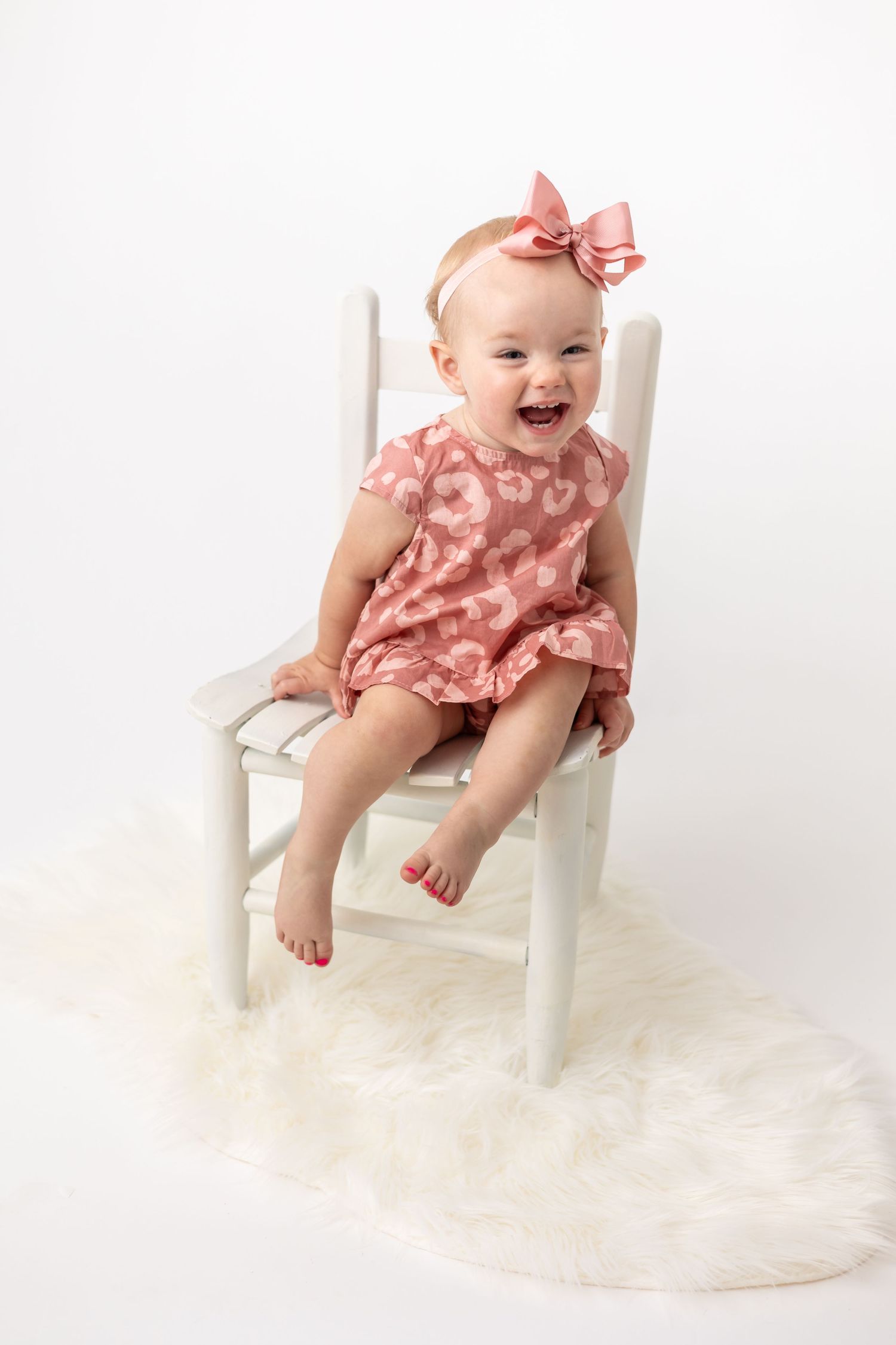 Happy baby wearing coral dress and bow sitting in white wooden chair on fuzzy rug having fun during her photography session.