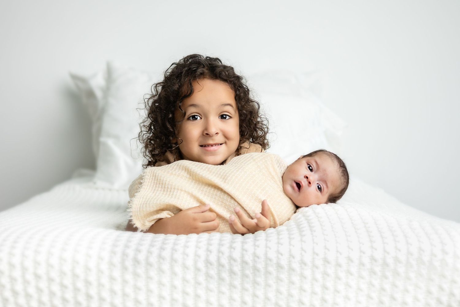 Toddler holding newborn baby sister on a white bed in Daleville, AL photography studio that specializes in dogs, babies, and families.