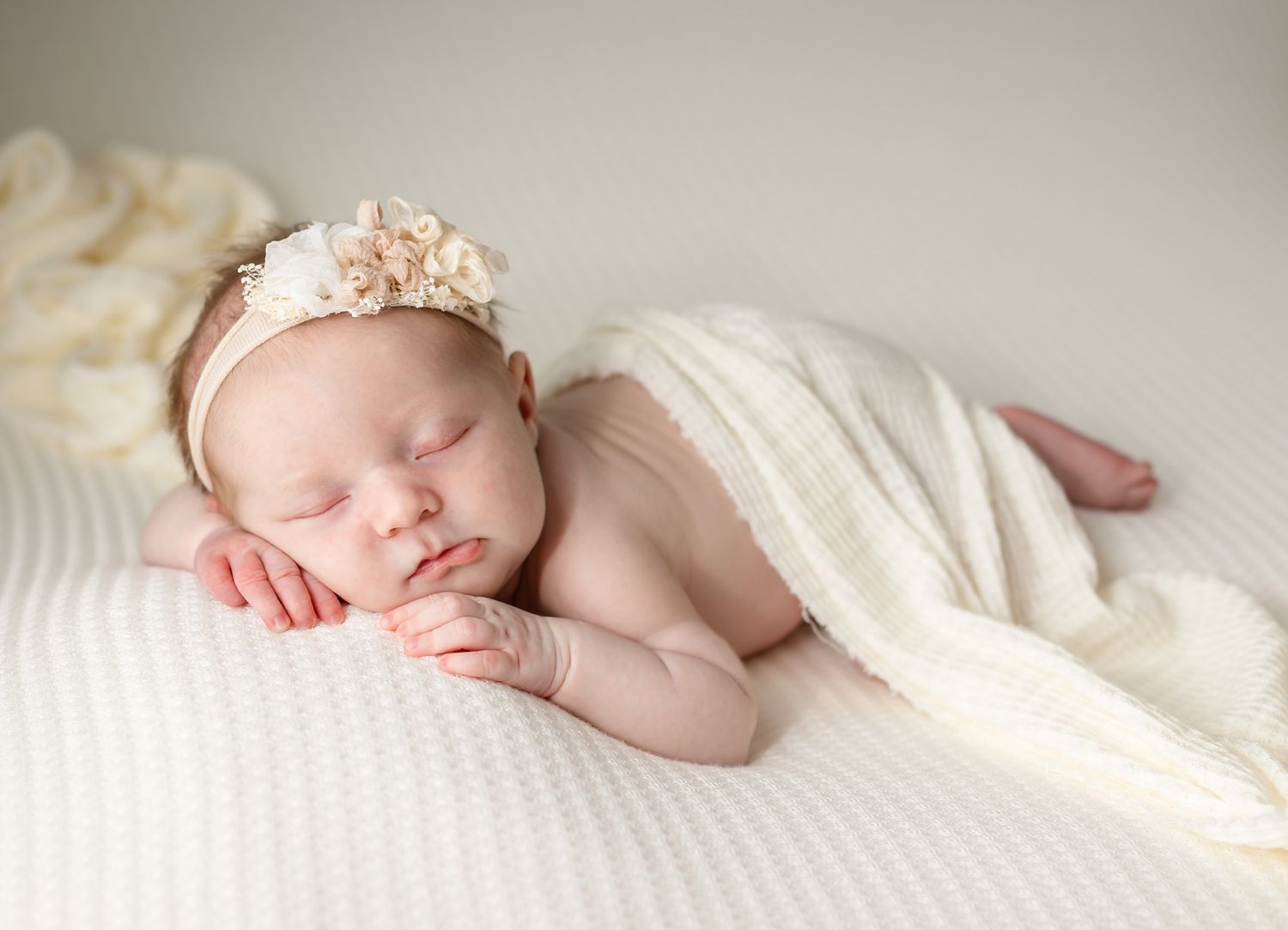 Sleeping newborn with pink headband partially covered by cream blanket.