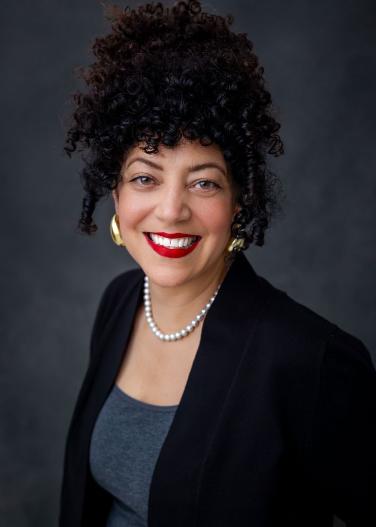 Professional headshot portrait featuring curly dark hair, pearl necklace, black blazer and bright red lipstick against dark background.