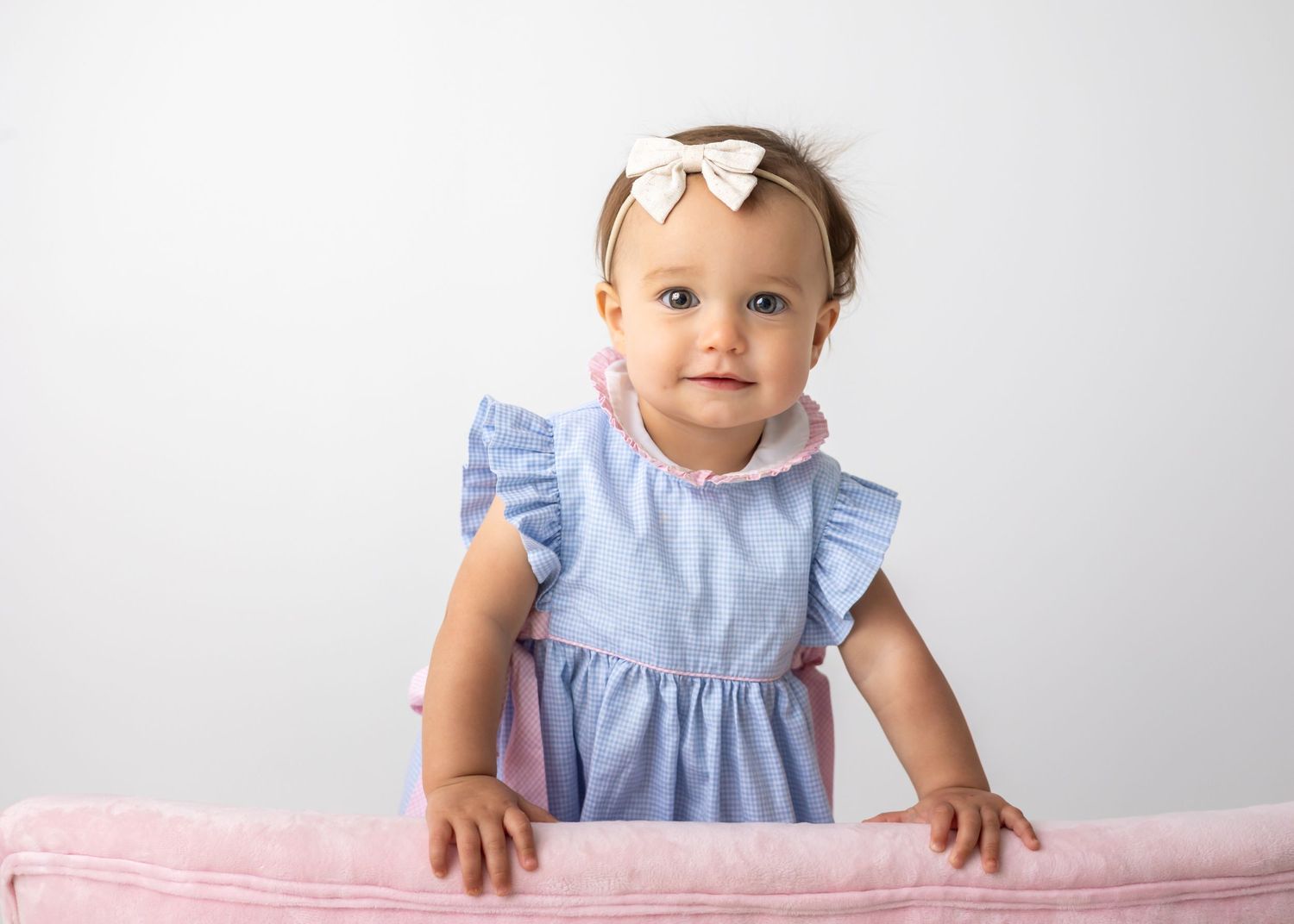 Adorable baby girl in blue ruffle dress and white bow headband standing at a pink blanket against white background.