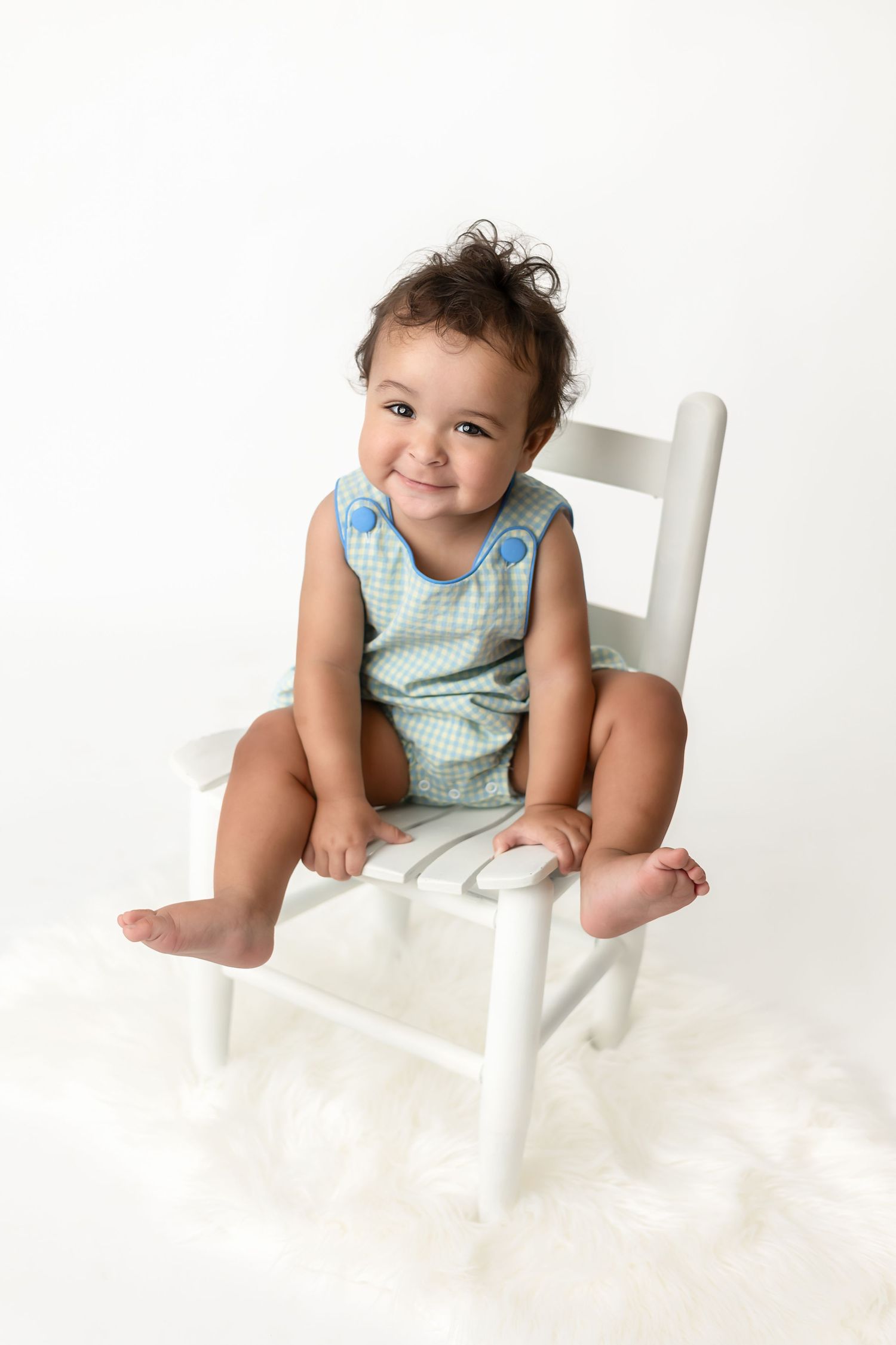 Baby in checkered outfit sits playfully on white chair against white background.
