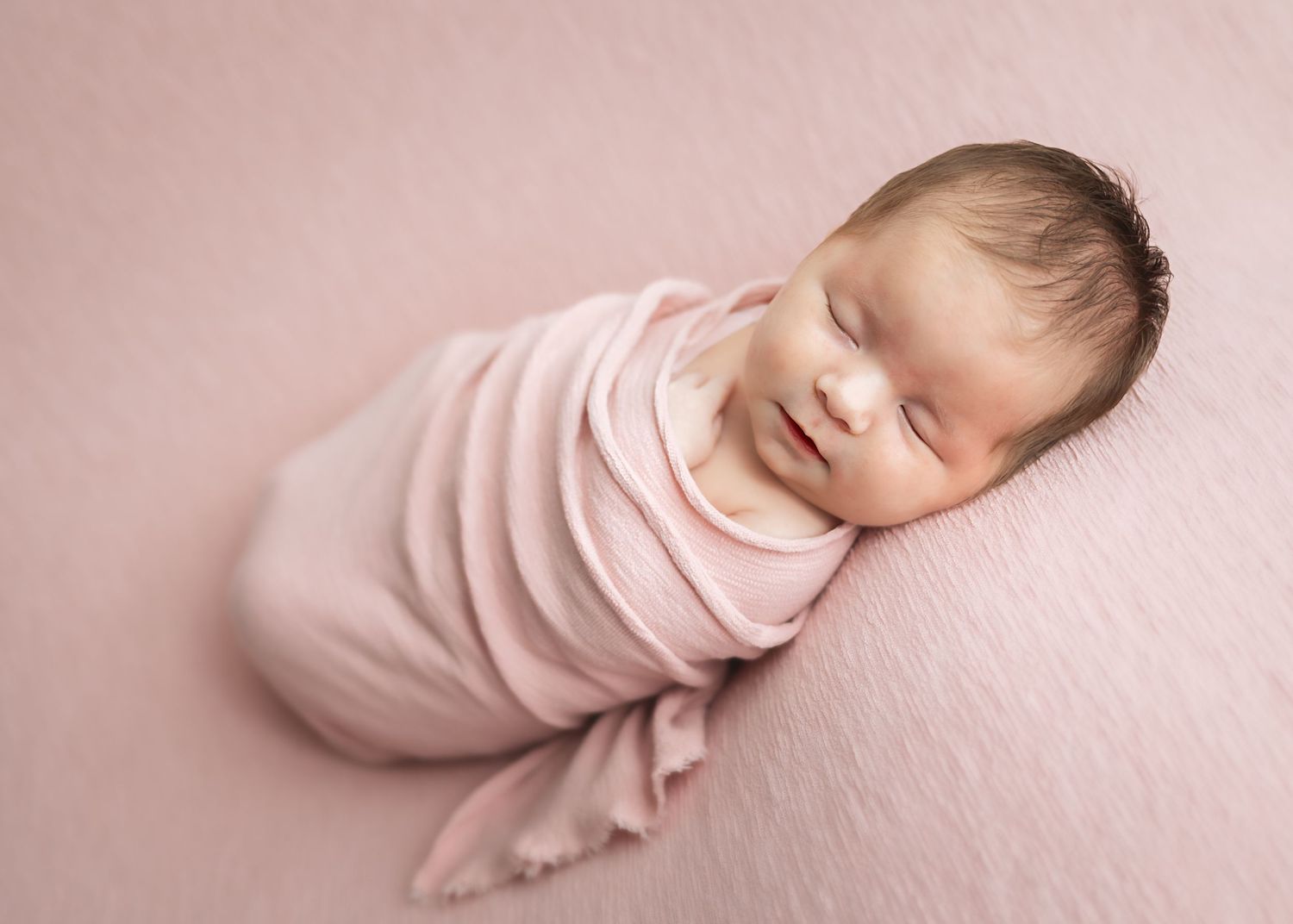 Sleeping newborn wrapped in soft pink swaddle on pink backdrop.