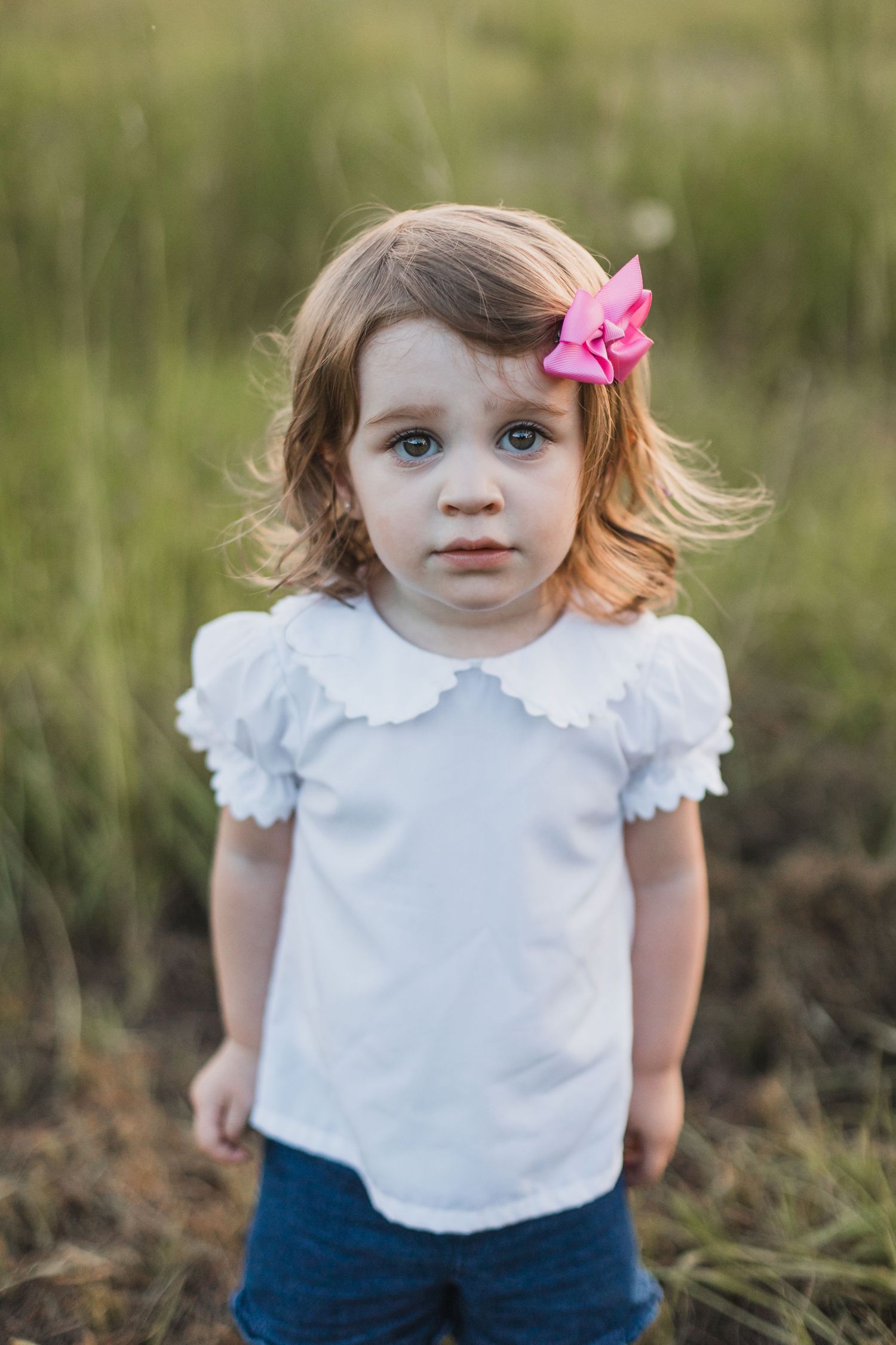 Young child in white ruffle shirt and denim with pink bow standing in tall grass during sunset.