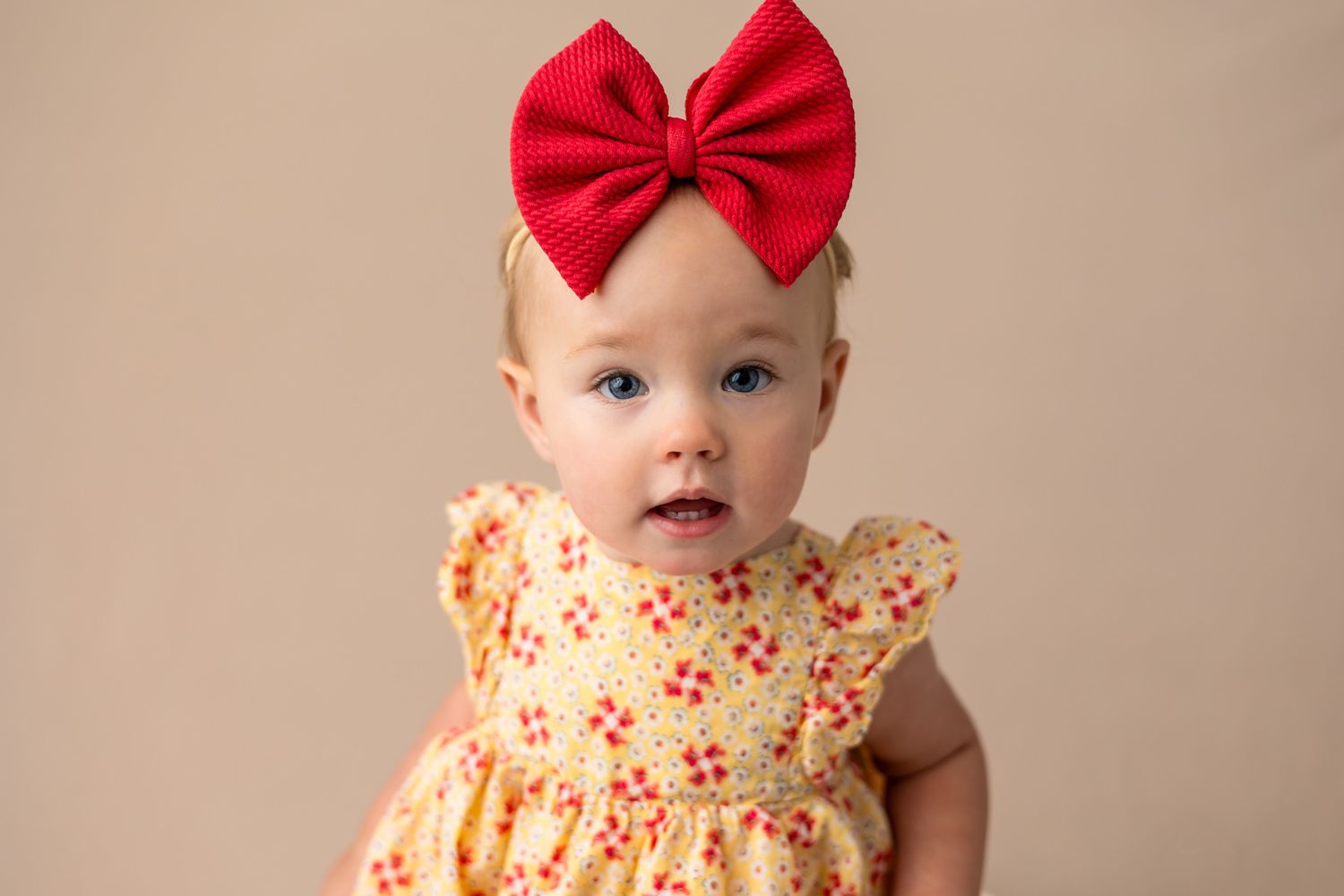 Baby wearing yellow floral dress and red bow headband against neutral background.