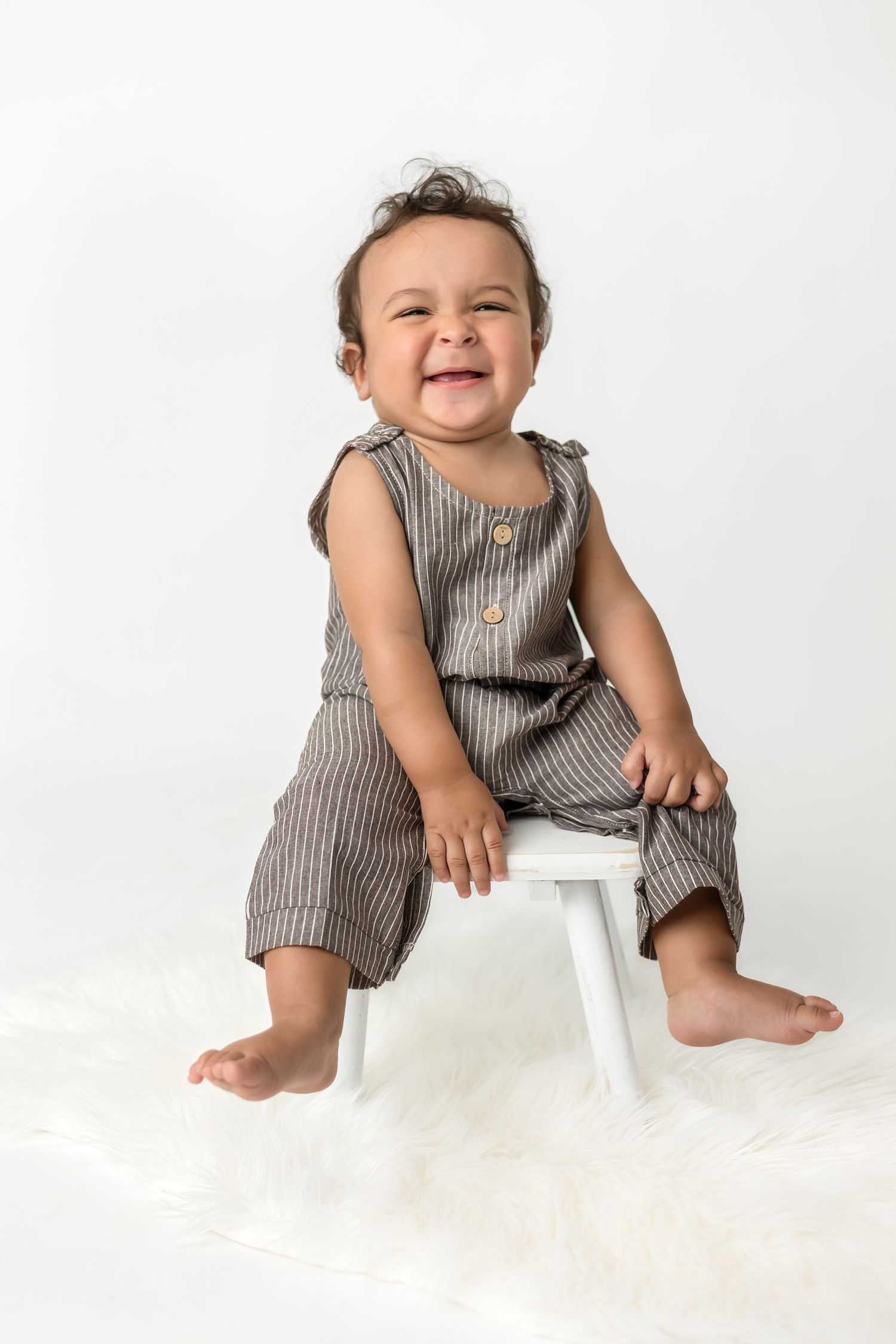Baby in striped romper sitting on white chair against light background.