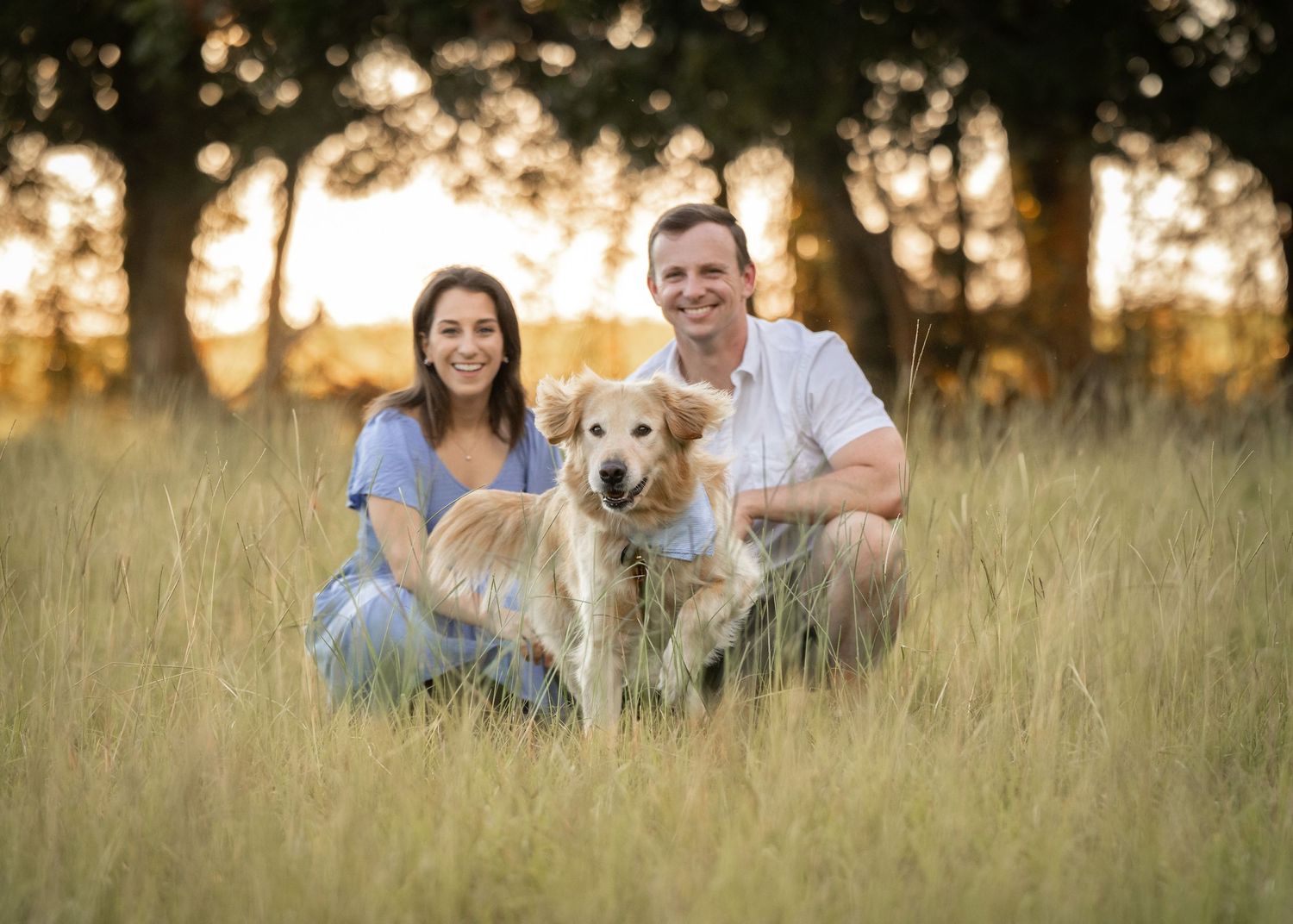 Family sitting together with their golden retriever in tall grass at sunset.