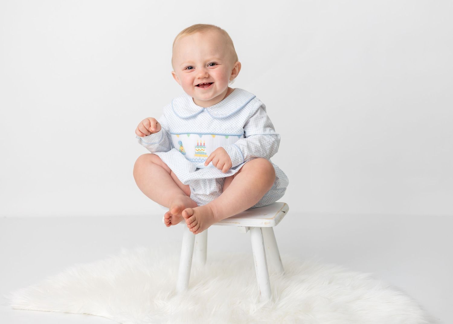 Smiling baby in light blue and white outfit sitting on small white chair.
