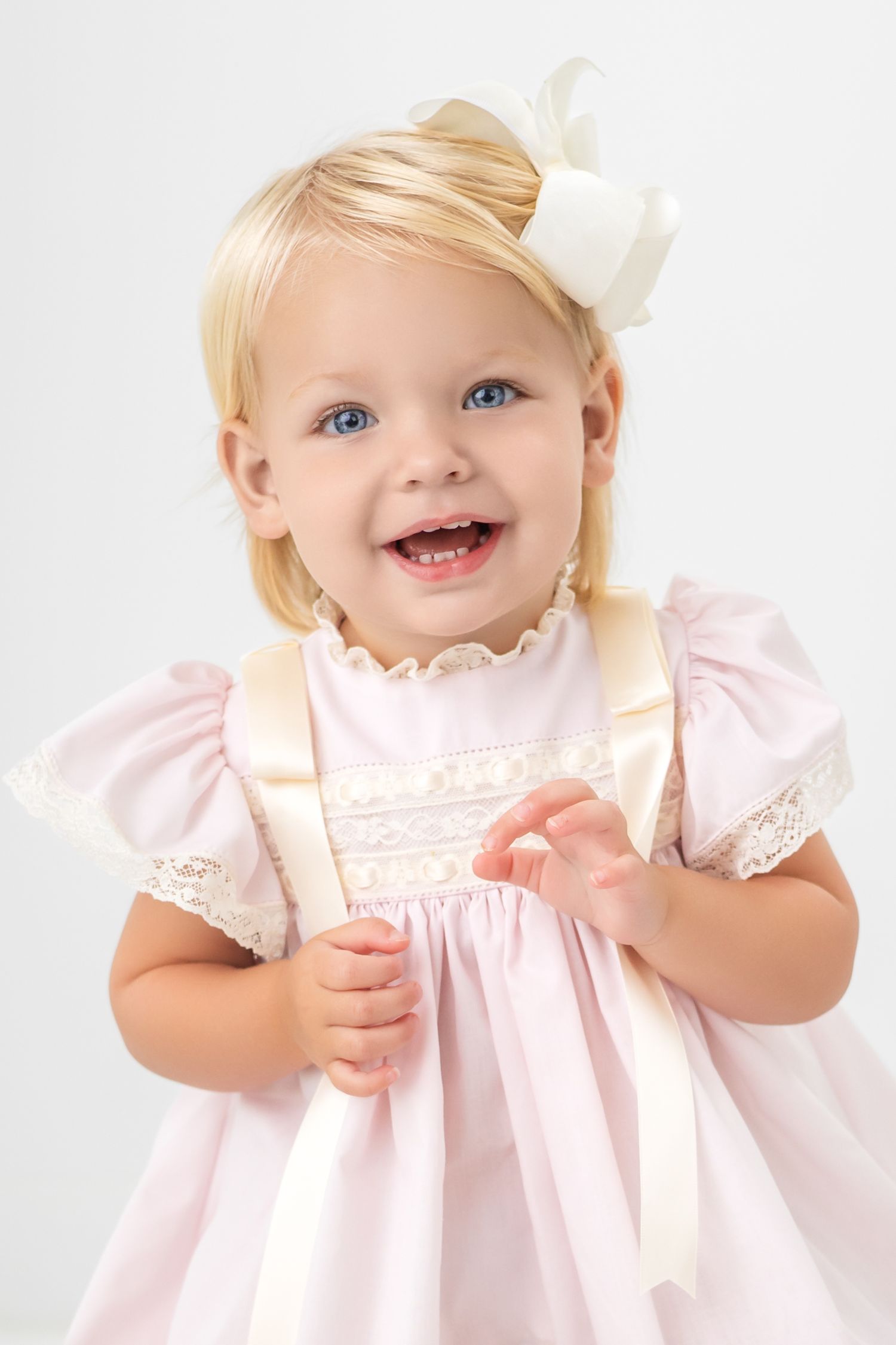 Smiling toddler girl with blue eyes and blonde hair wearing a pink dress with lace trim and white bow headband.