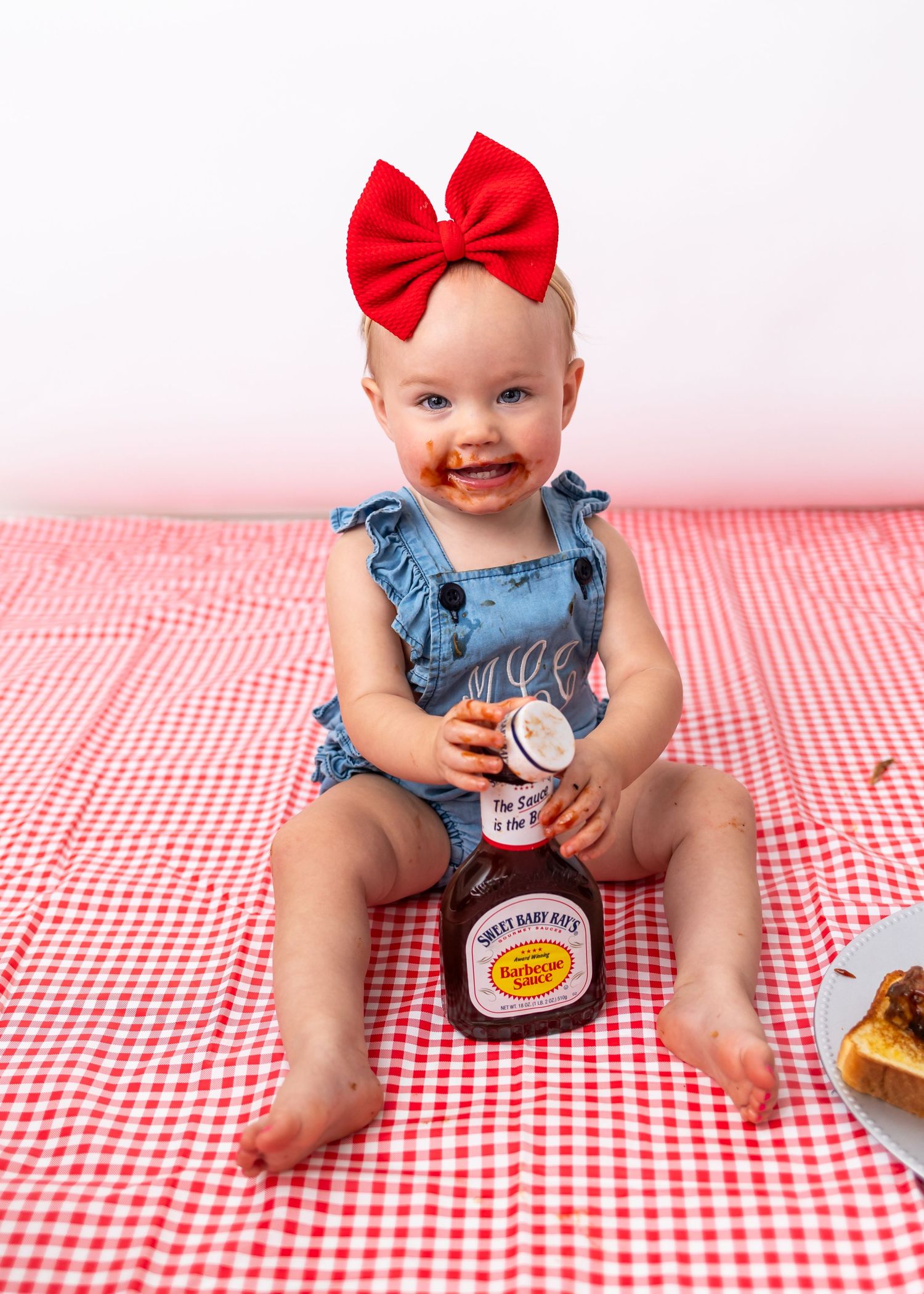 Baby in denim outfit playing with BBQ sauce bottles on red and white checkered tablecloth.