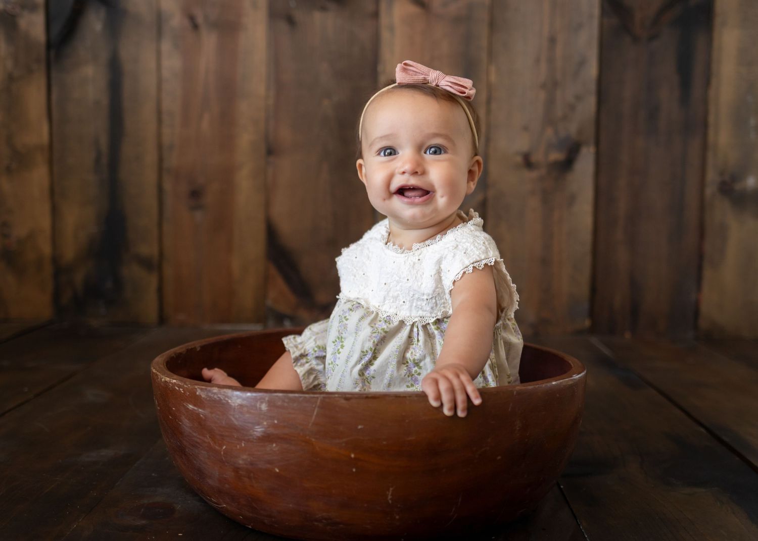 Baby sitting in wooden bowl wearing bow and lace outfit.