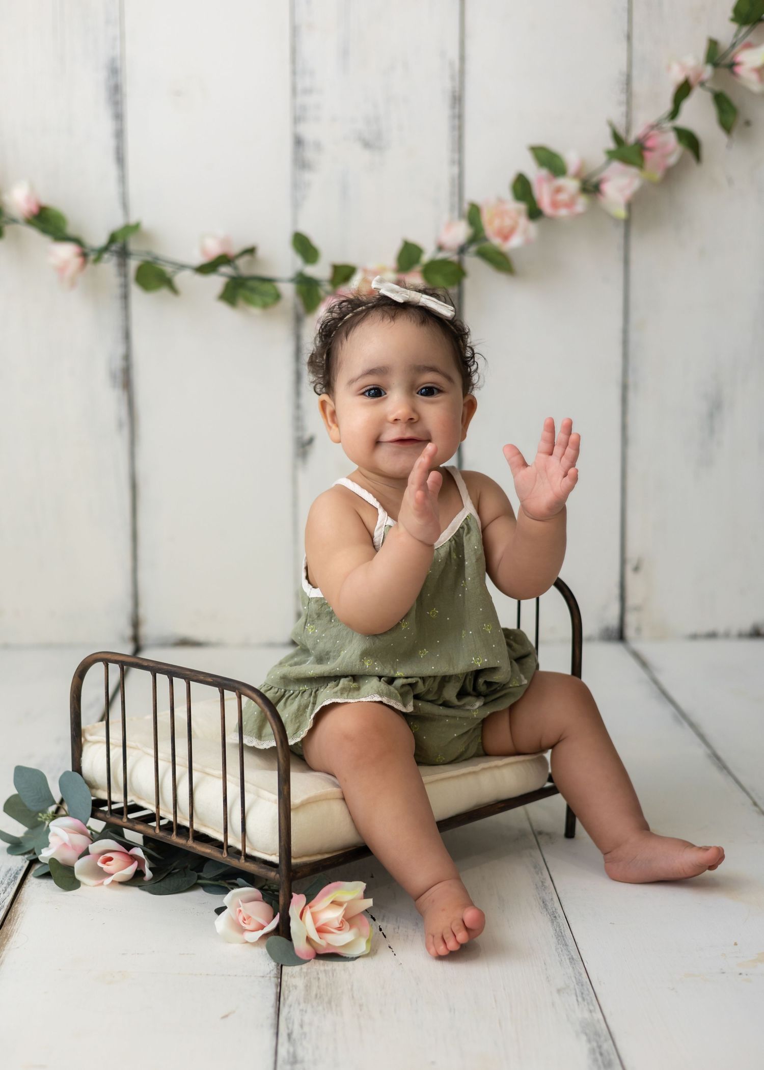Smiling baby girl in green romper sitting on miniature iron bed with pink floral garland backdrop in professional photo.