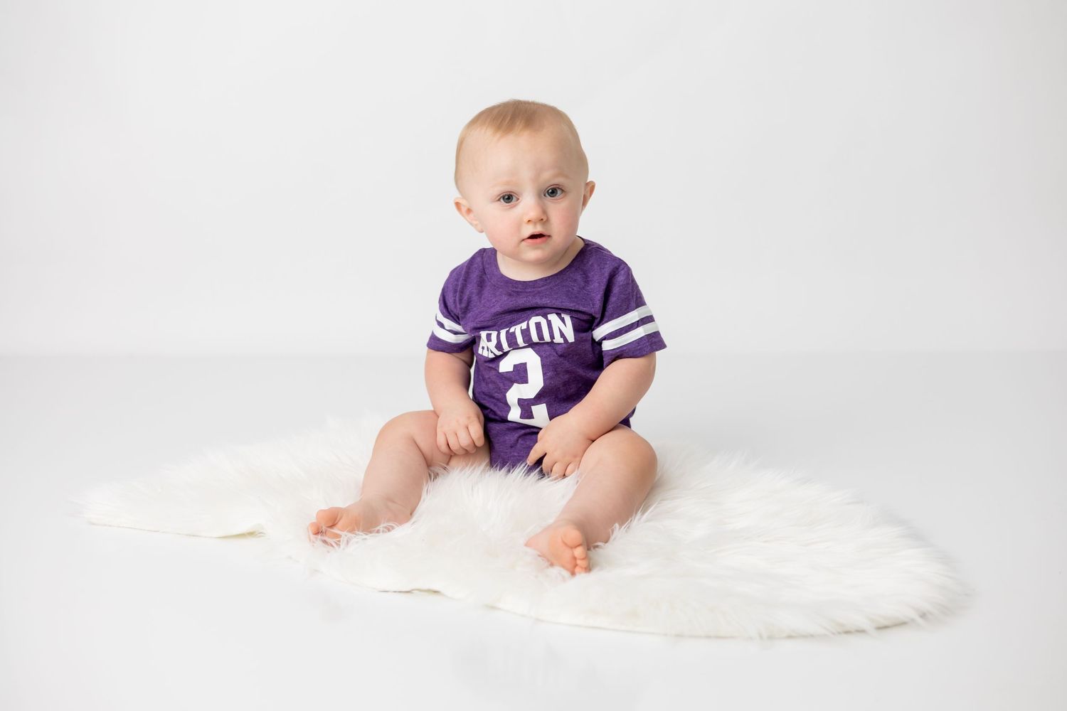 Baby wearing purple sports jersey sitting on fluffy white rug.