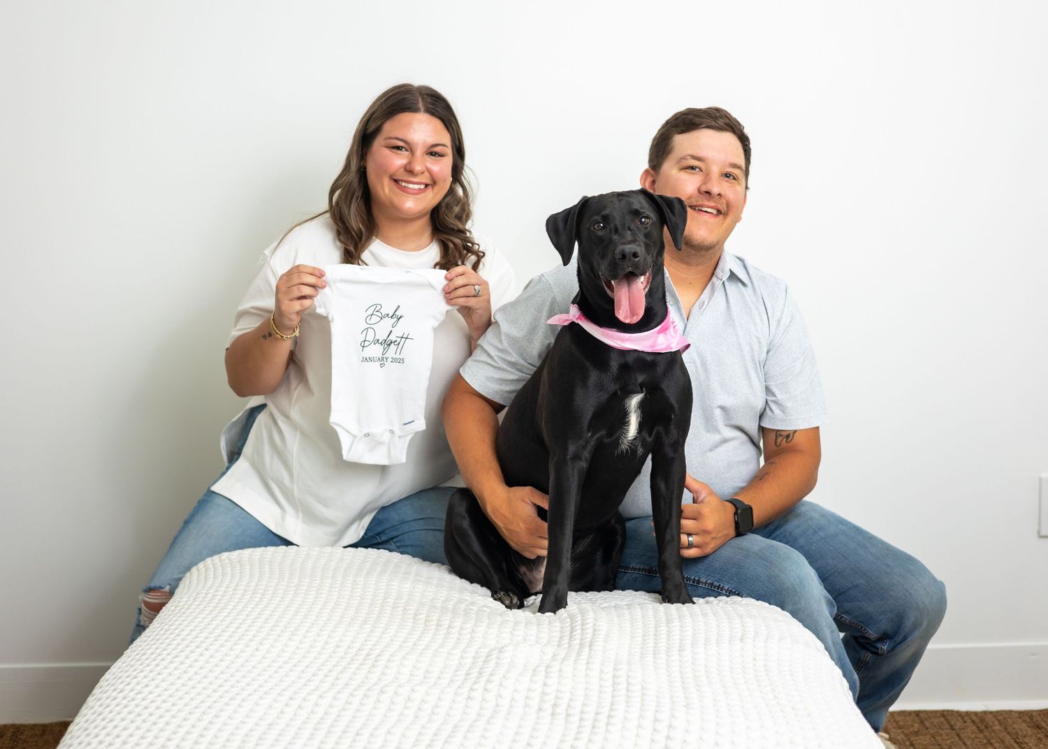 Couple relaxing on white mattress with their black dog for casual indoor photos.