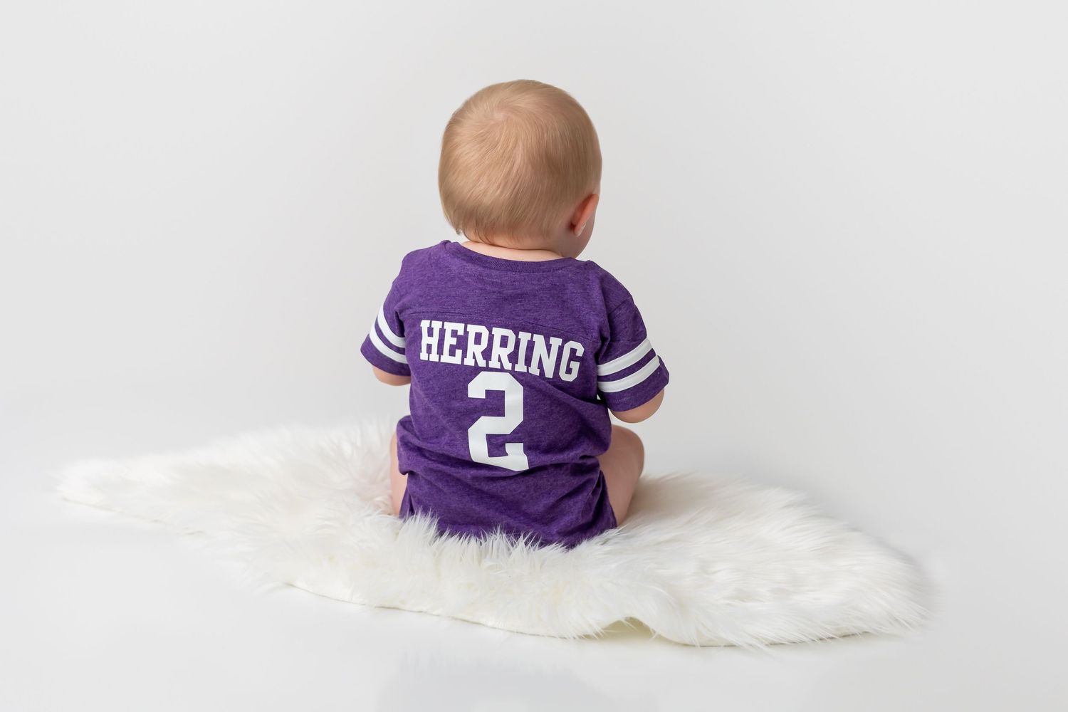Back view of baby wearing purple sports jersey with number 2 sitting on white rug.