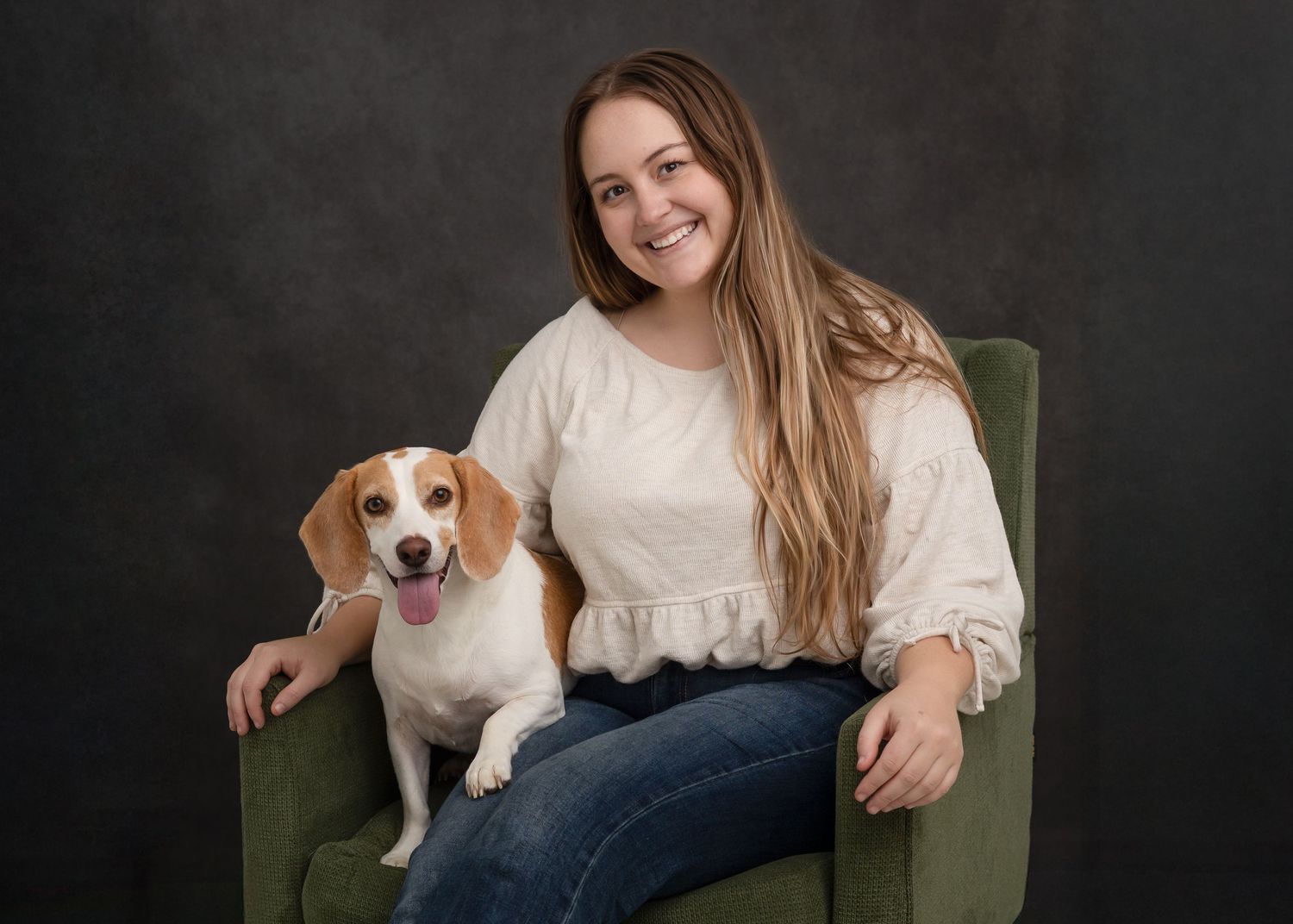 A beagle puppy sits on a green velvet chair with someone in jeans and a white top.