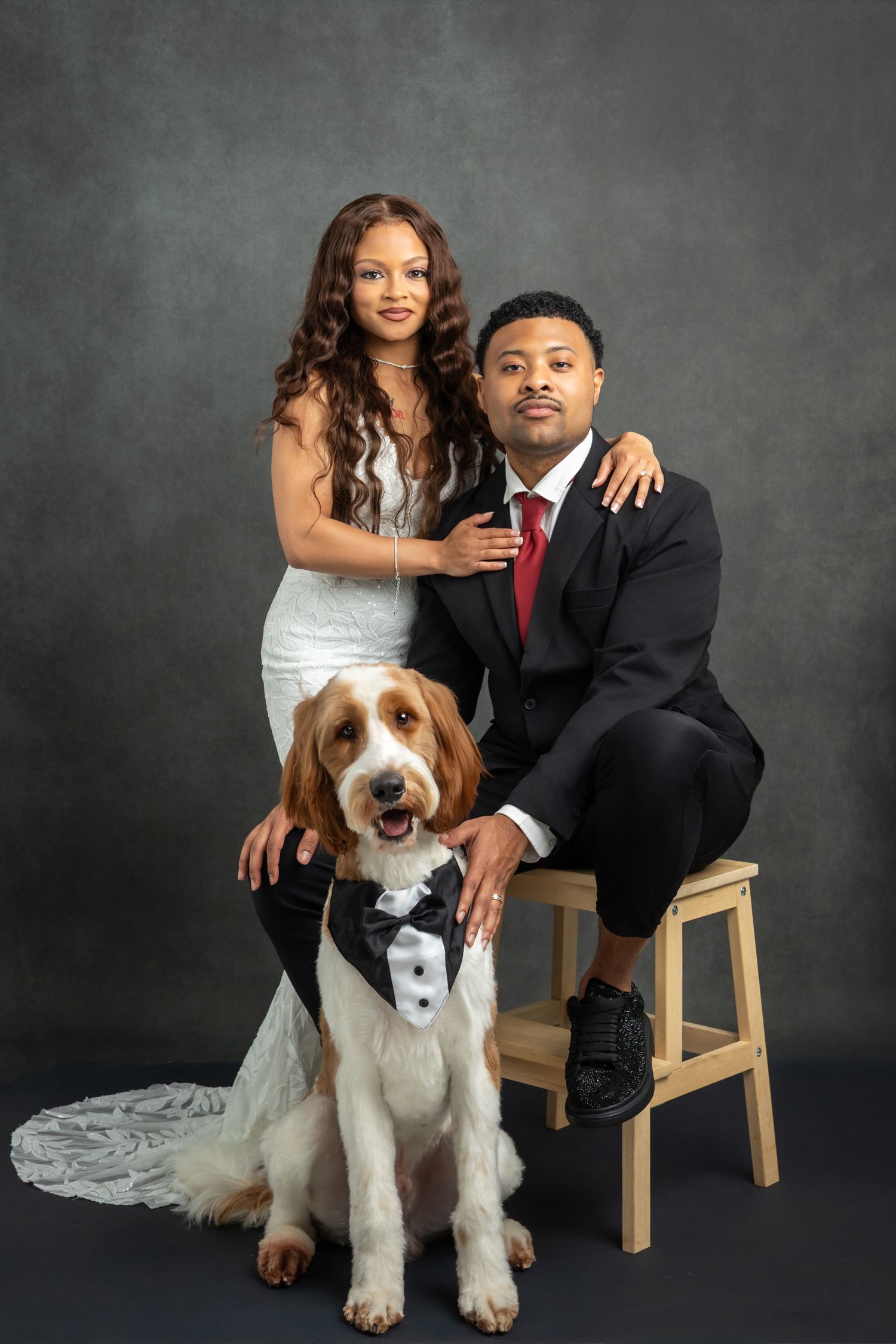 Couple in wedding attire poses with their dog wearing a tuxedo for a formal studio portrait photo.