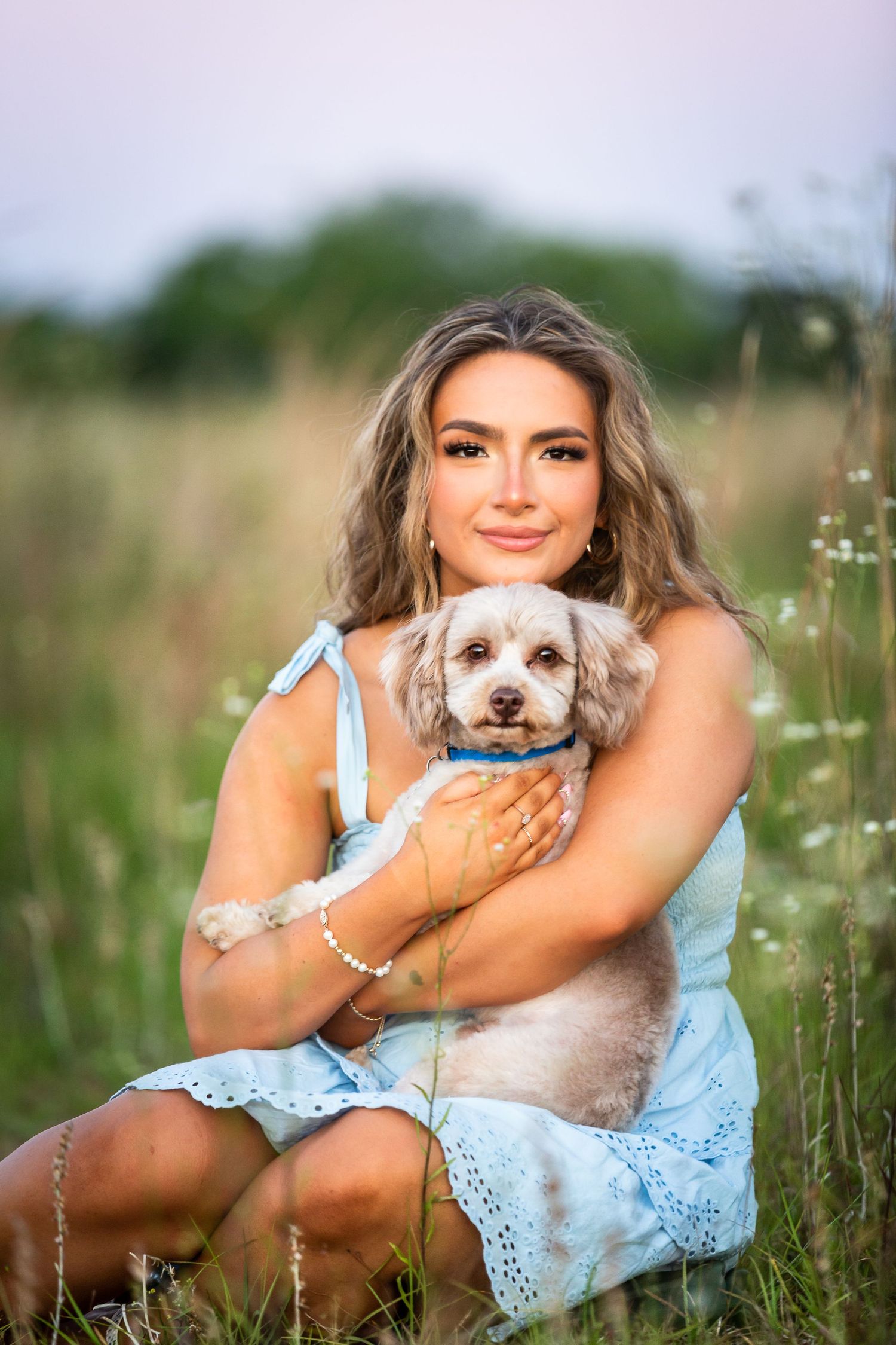 Woman in a light blue dress sits in a wildflower meadow holding a fluffy white and brown dog during a summer photoshoot.
