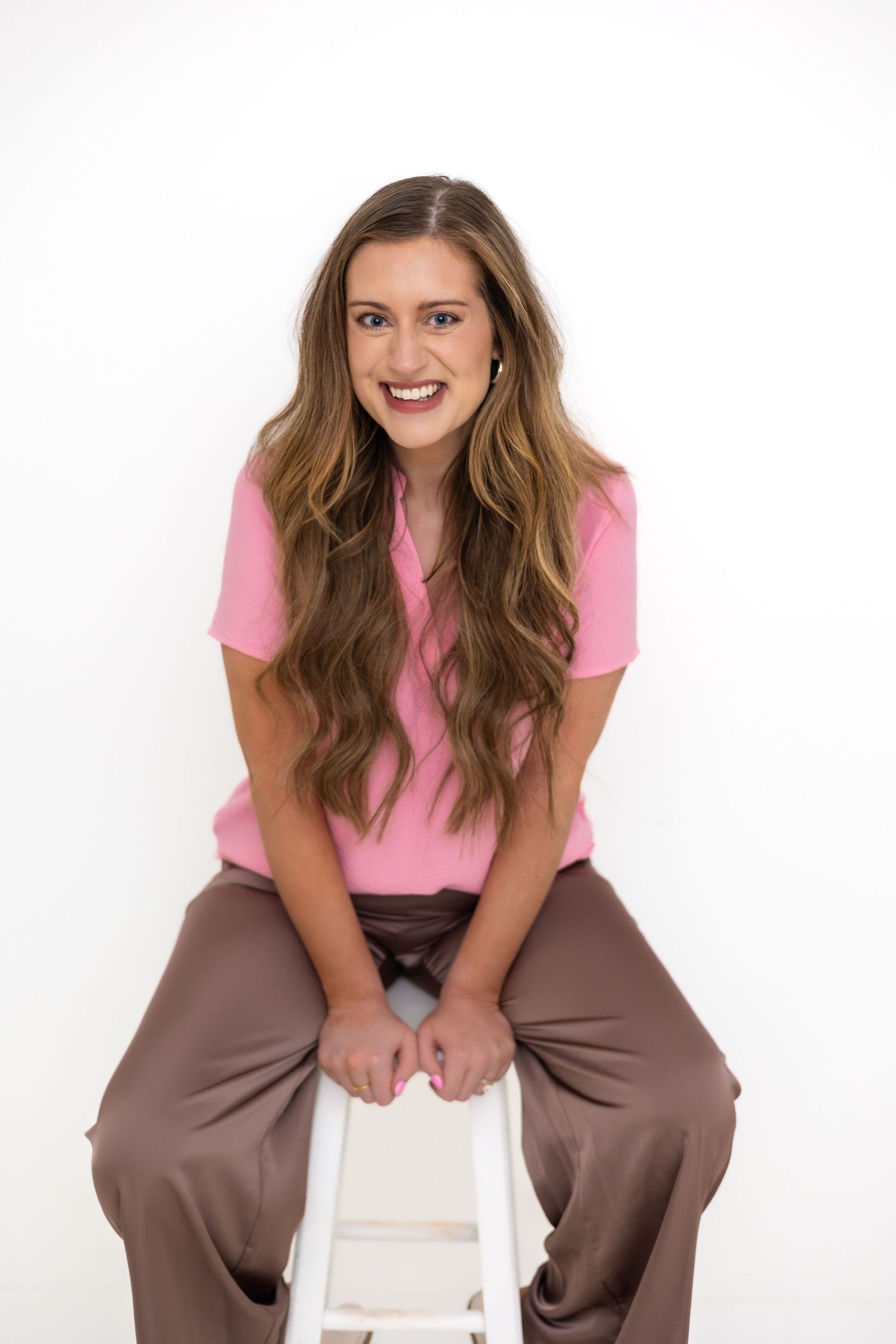 A person wearing a pink t-shirt and brown pants sits and smiles on a white chair against a bright background.