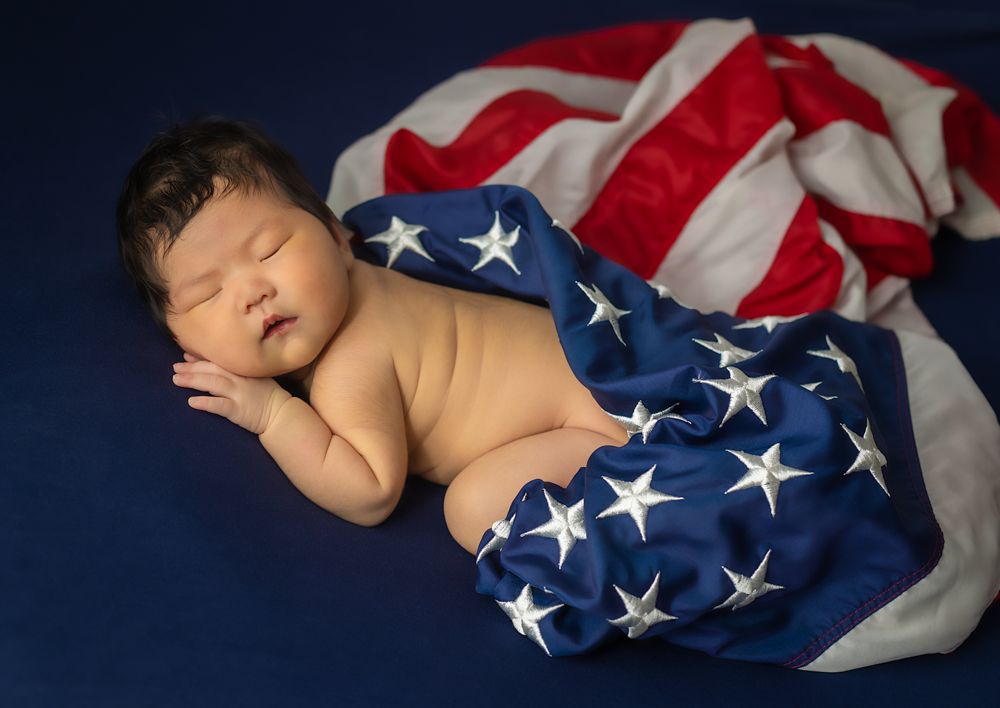 Sleeping newborn wrapped in an American flag on dark blue background.