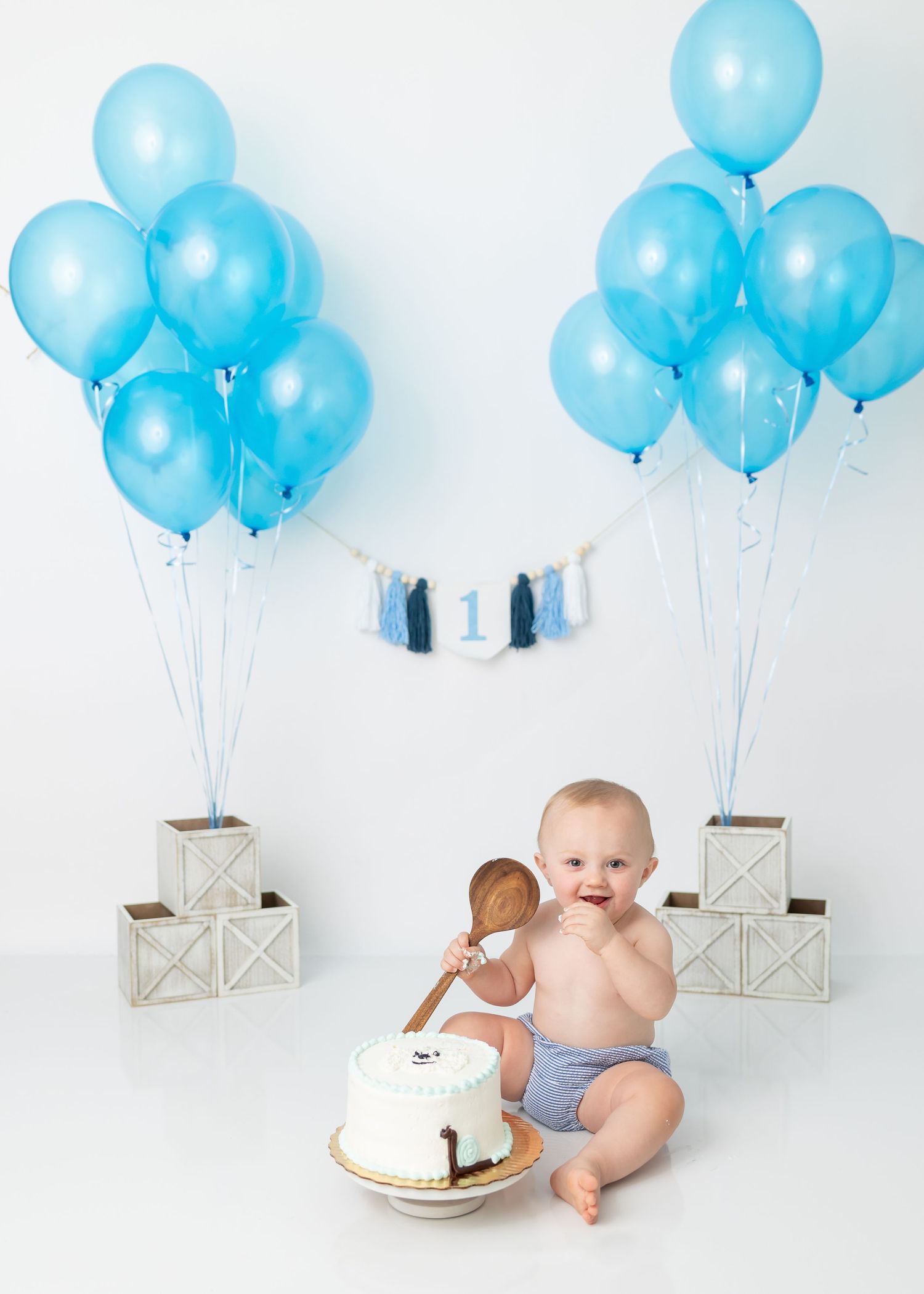 Baby in diaper enjoys cake smash photo session with blue balloons and wooden crates.