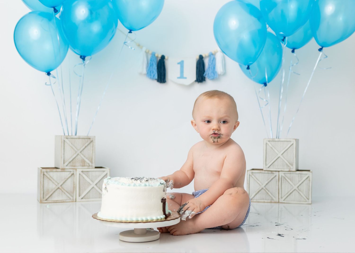Baby cake smash photo with blue balloons and white boxes against light background.
