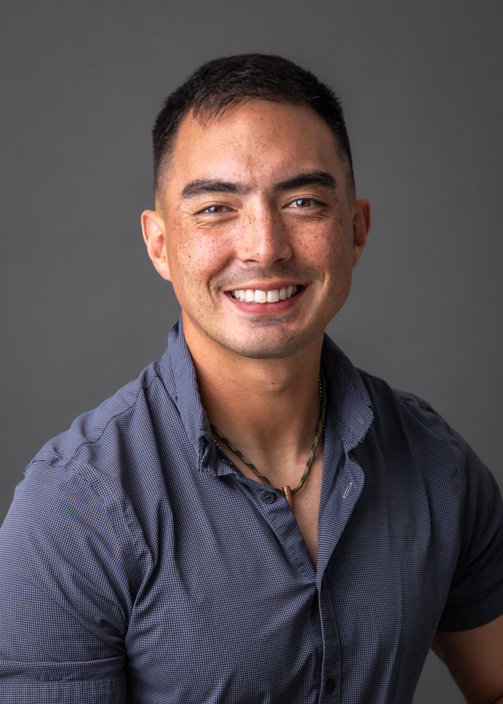 Professional headshot portrait of a person in a dark button-up shirt smiling warmly against a gray background.