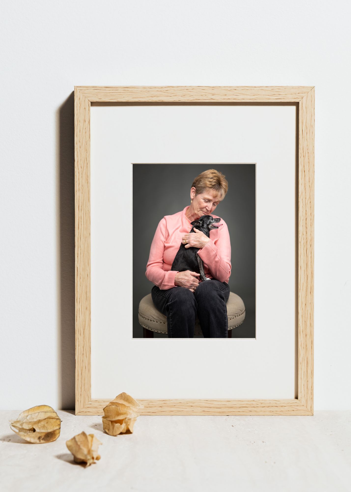Three framed photographs of someone in a pink sweater holding a small black cat are displayed with seashells on a white wall.