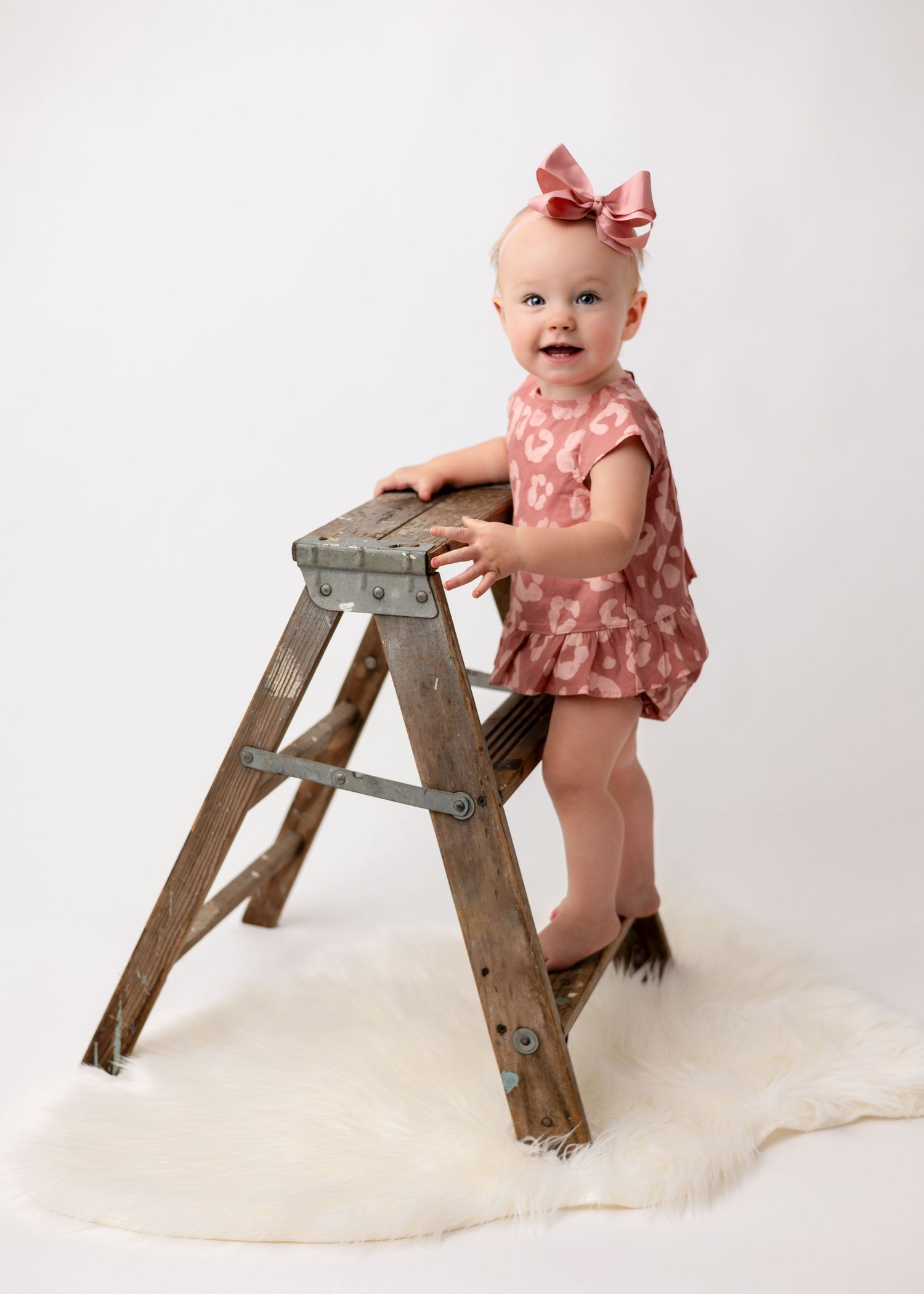A baby in a pink ruffled outfit plays with a vintage wooden stepladder against a white backdrop.