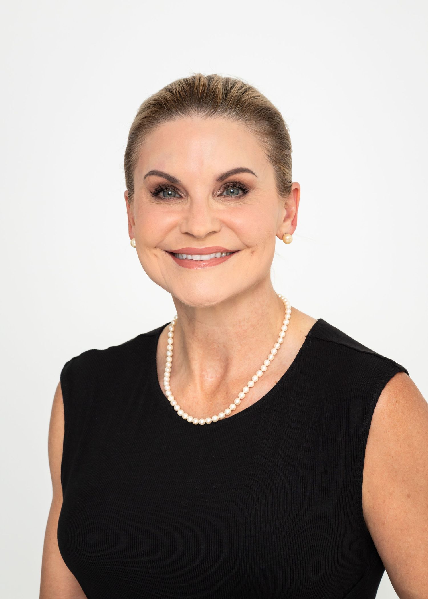 A person in a black sleeveless top and pearl necklace smiles brightly against a white background.